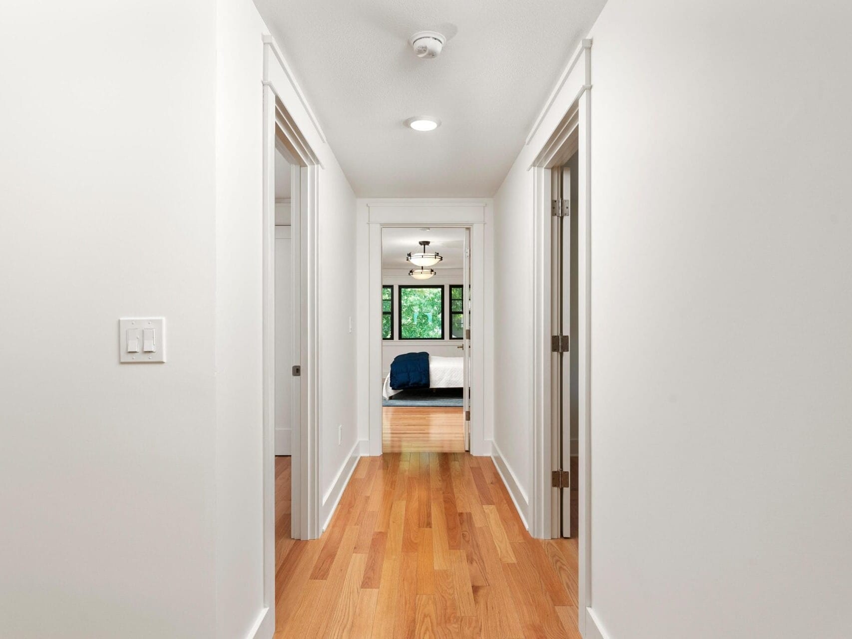 A bright hallway with wooden floors and white walls leads to a bedroom at the end. The bedroom features a bed with dark bedding, two side tables, and a window with a view of greenery. Recessed lighting and a smoke detector are on the ceiling.