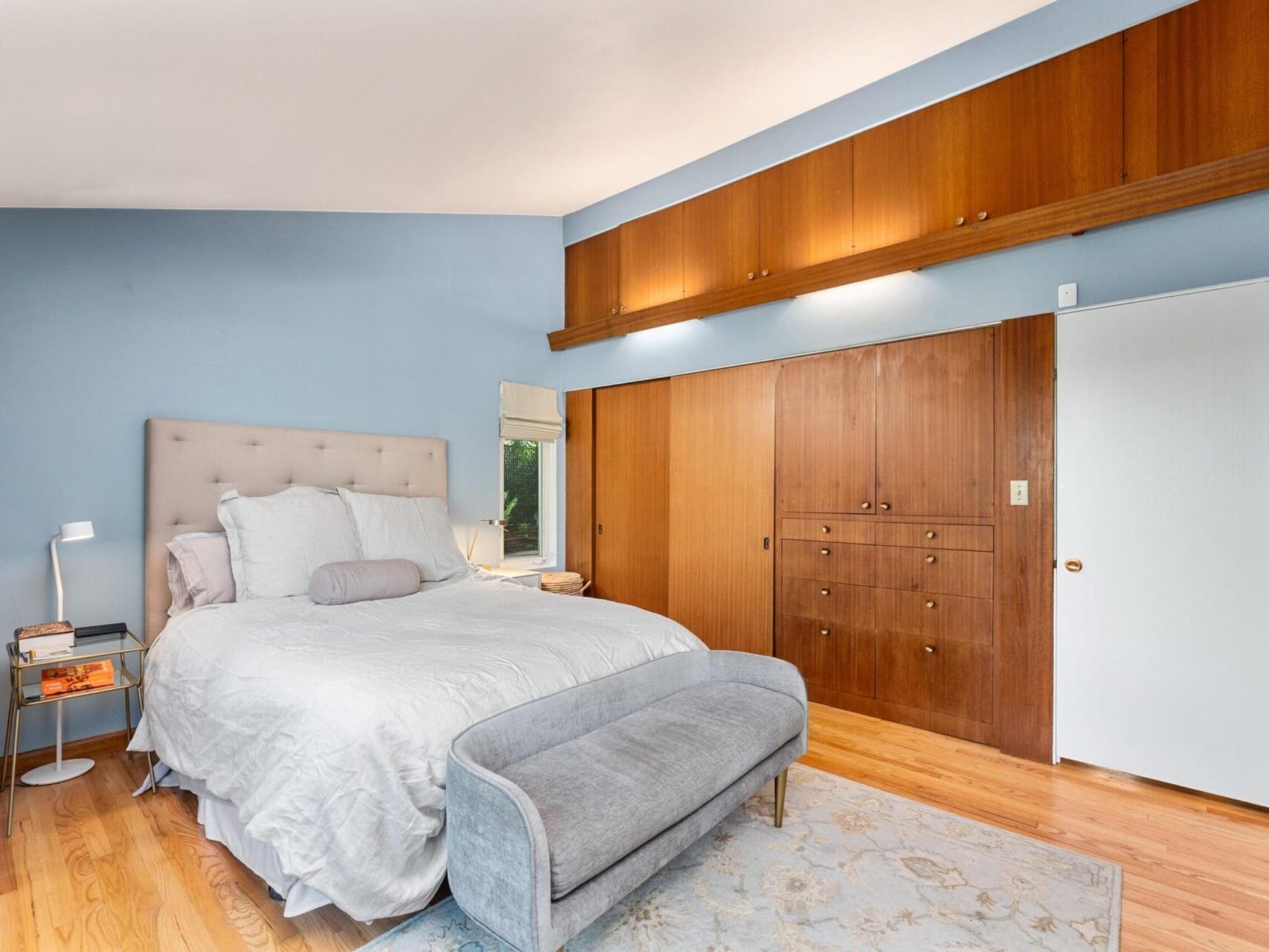 A serene bedroom with light blue walls features a bed with a tufted headboard, a gray bench, and wooden cabinets along the wall. A stack of books sits beside the bed on a modern lampstand, adding a cozy touch to the room.