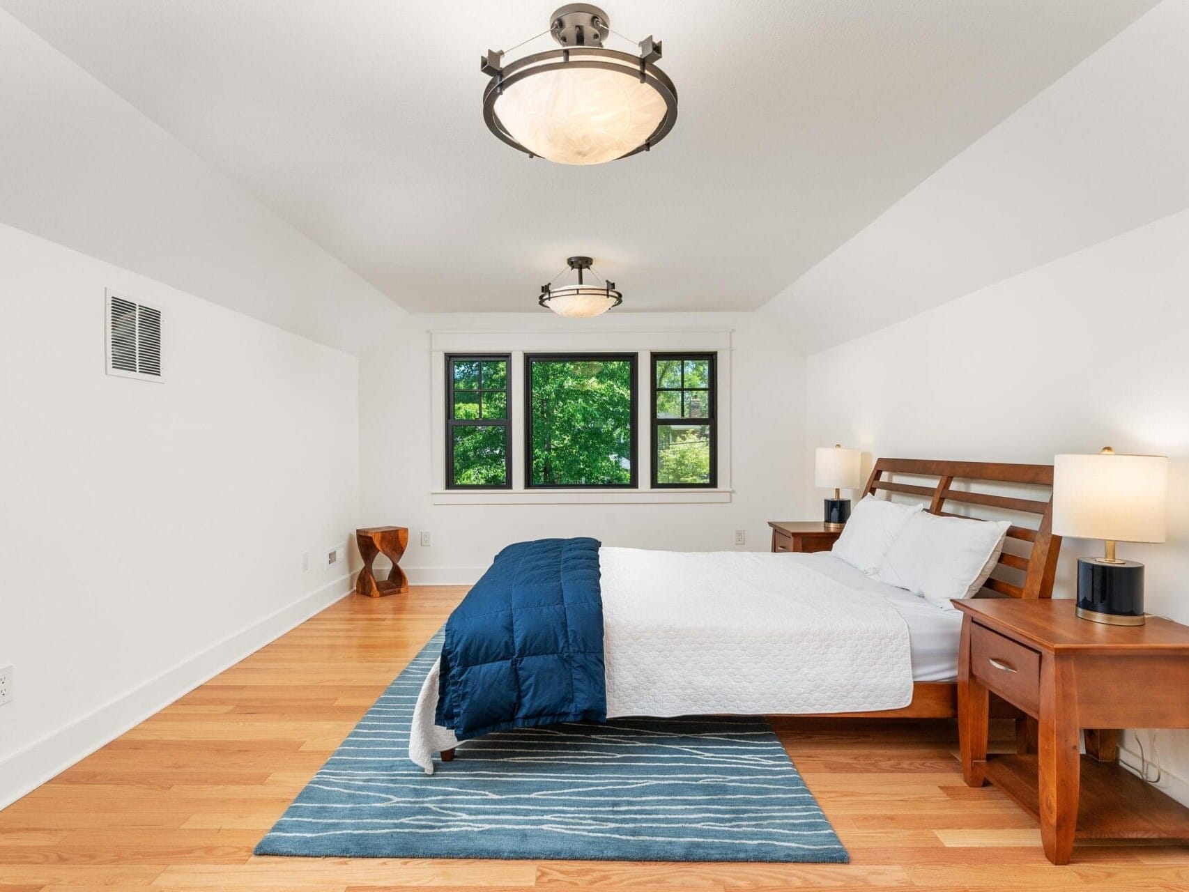 A minimalist bedroom with a wooden bed frame, blue bedding, and white pillows. Two wooden nightstands with lamps flank the bed. A window with a view of greenery is in the background, and a striped rug lies on a light wooden floor.