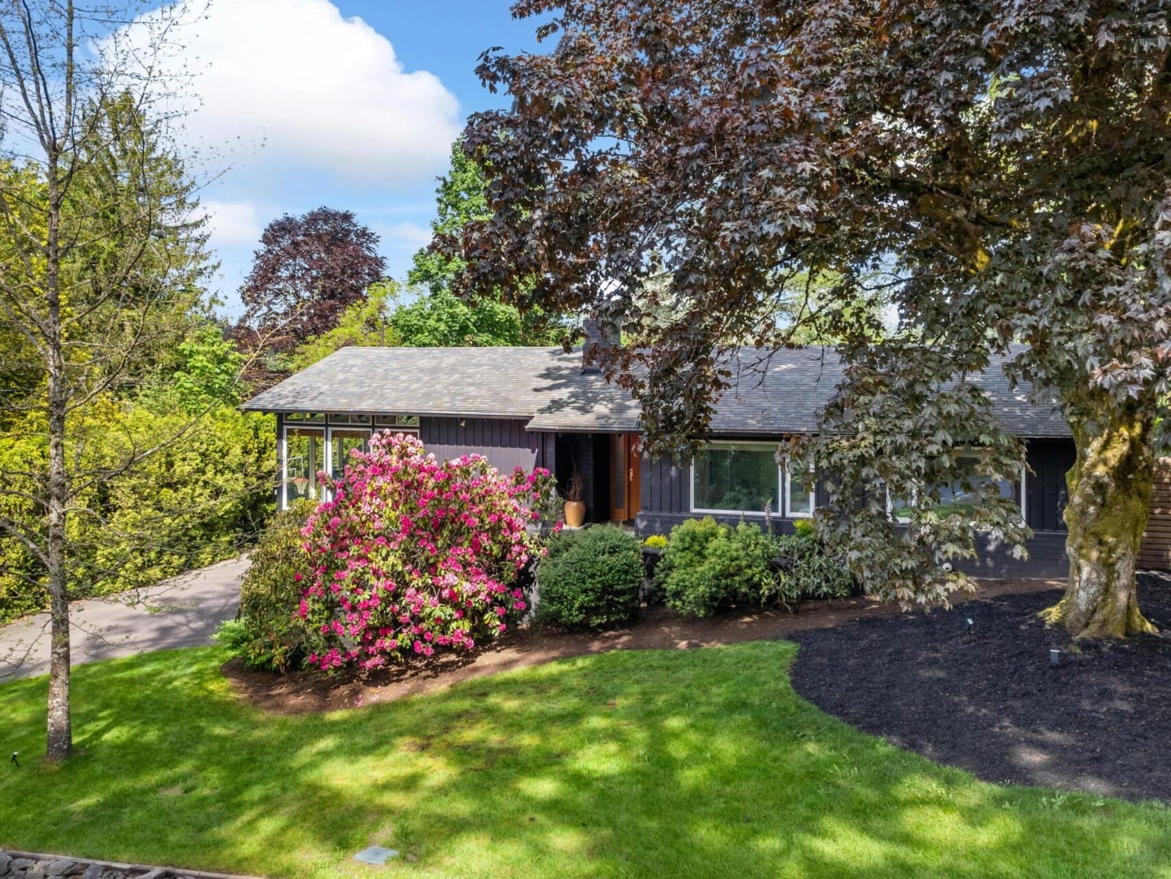 A single-story house surrounded by lush greenery, with a large flowering bush near the entrance. The front yard features a well-maintained lawn, trees, and shrubs, under a clear blue sky.