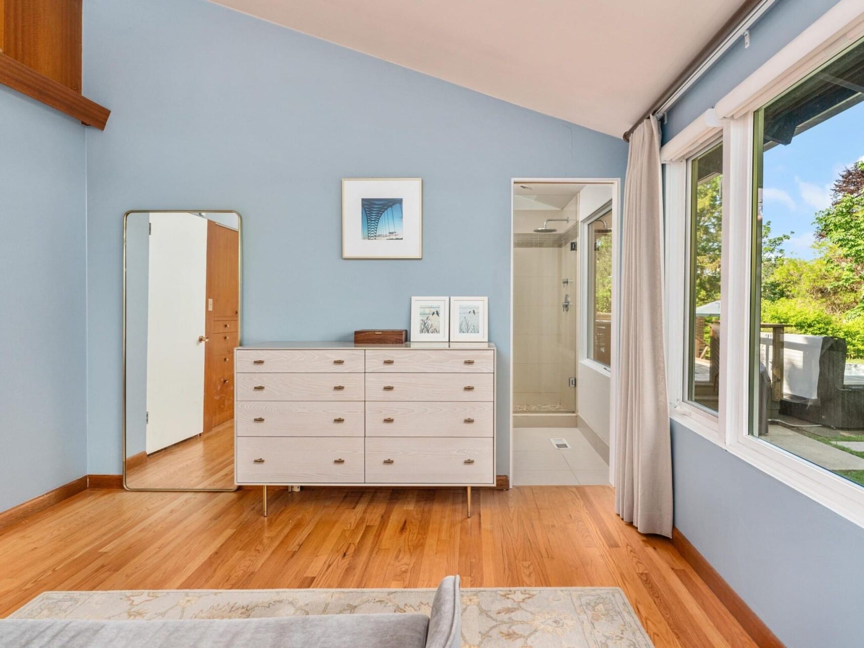 A bright bedroom with light blue walls, featuring a large mirror, a white dresser with framed artwork, and a window showing a garden outside. Wood flooring and a plush rug add warmth to the minimalist space.