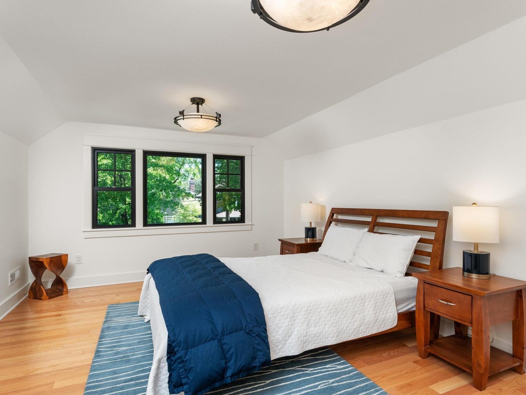 A cozy bedroom with a wooden bed frame, white bedding, and a dark blue throw blanket. Two bedside tables with lamps flank the bed. Large windows with black frames provide natural light. A striped rug covers the wooden floor.