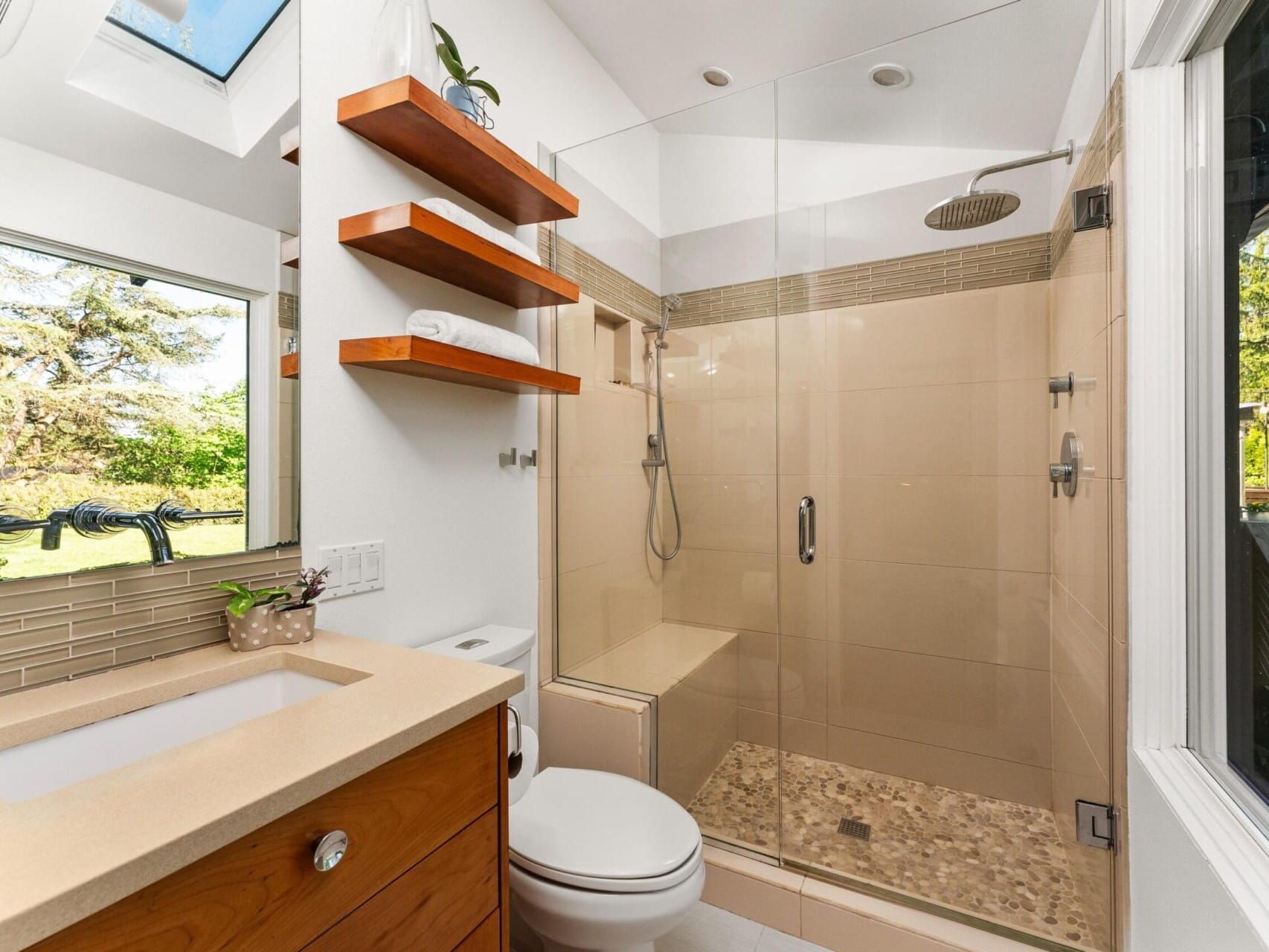 Modern bathroom with a glass-enclosed shower featuring a rainfall showerhead. There is a wooden vanity with a sink, a small plant, and shelving with towels. A large window overlooks a leafy backyard, and a skylight lets in additional natural light.