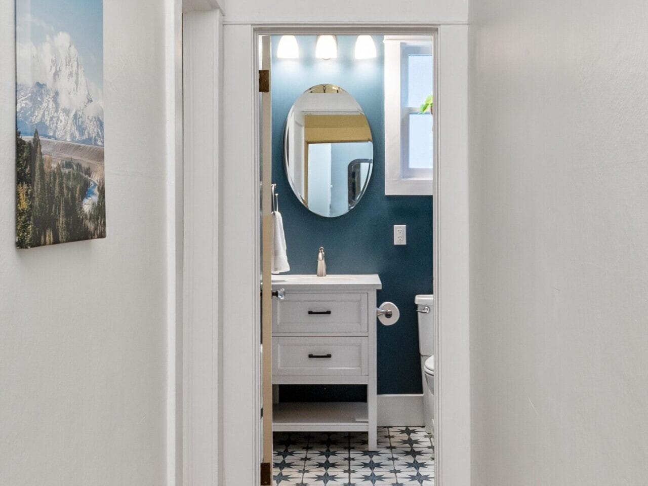 A narrow hallway with white walls leading to a bathroom. The bathroom features blue walls, a round mirror, and a white vanity. A colorful orange rug lies on the wooden floor, and a scenic landscape photo hangs on the left wall.