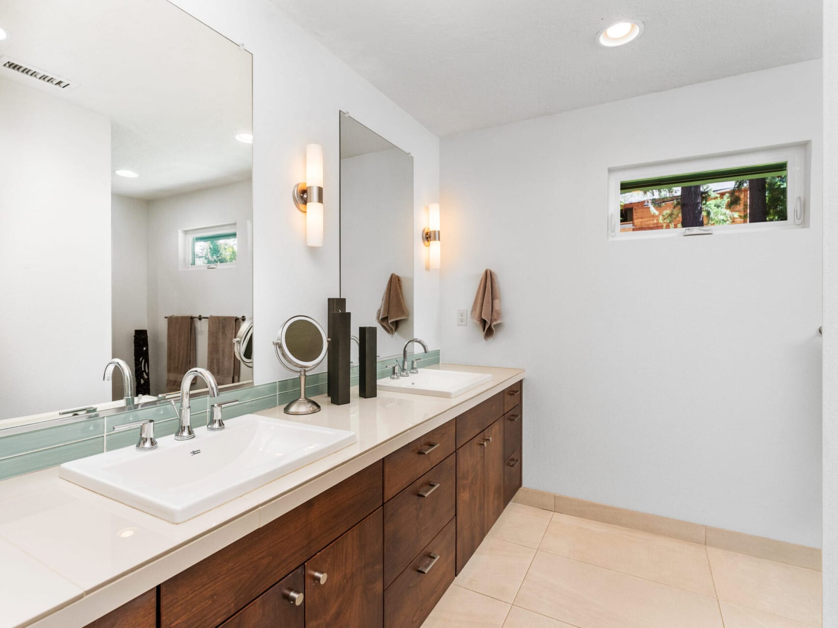 A modern bathroom featuring a double-sink vanity with dark wood cabinets and a green-tiled backsplash. Two mirrors are mounted above the sinks, with wall sconces providing lighting. A small window allows natural light to enter the space.