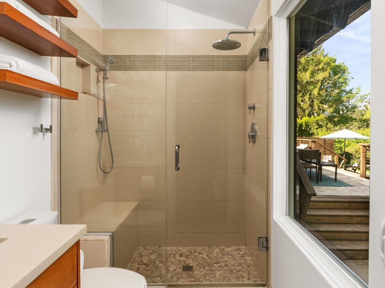 Modern bathroom with glass-enclosed shower, beige tiles, and pebble floor. Wooden shelves hold white towels above a toilet. A large window offers a view of a deck and trees outside. Skylight brings natural light into the space.