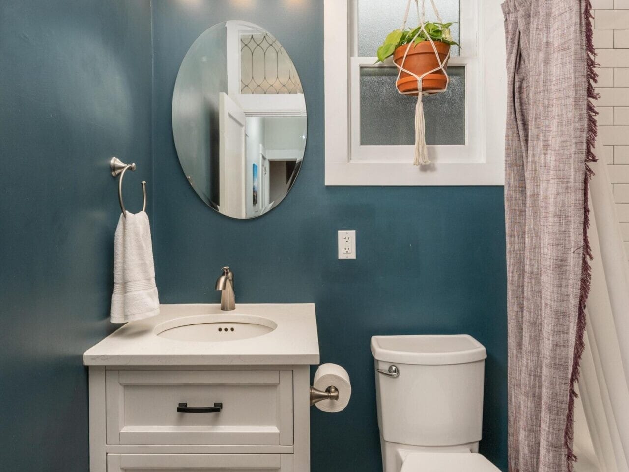 A bathroom with teal walls, a round mirror above a white vanity, and a white toilet. Theres a hanging potted plant near a small window. The floor has a geometric tile pattern, and a shower curtain hangs beside the tub.