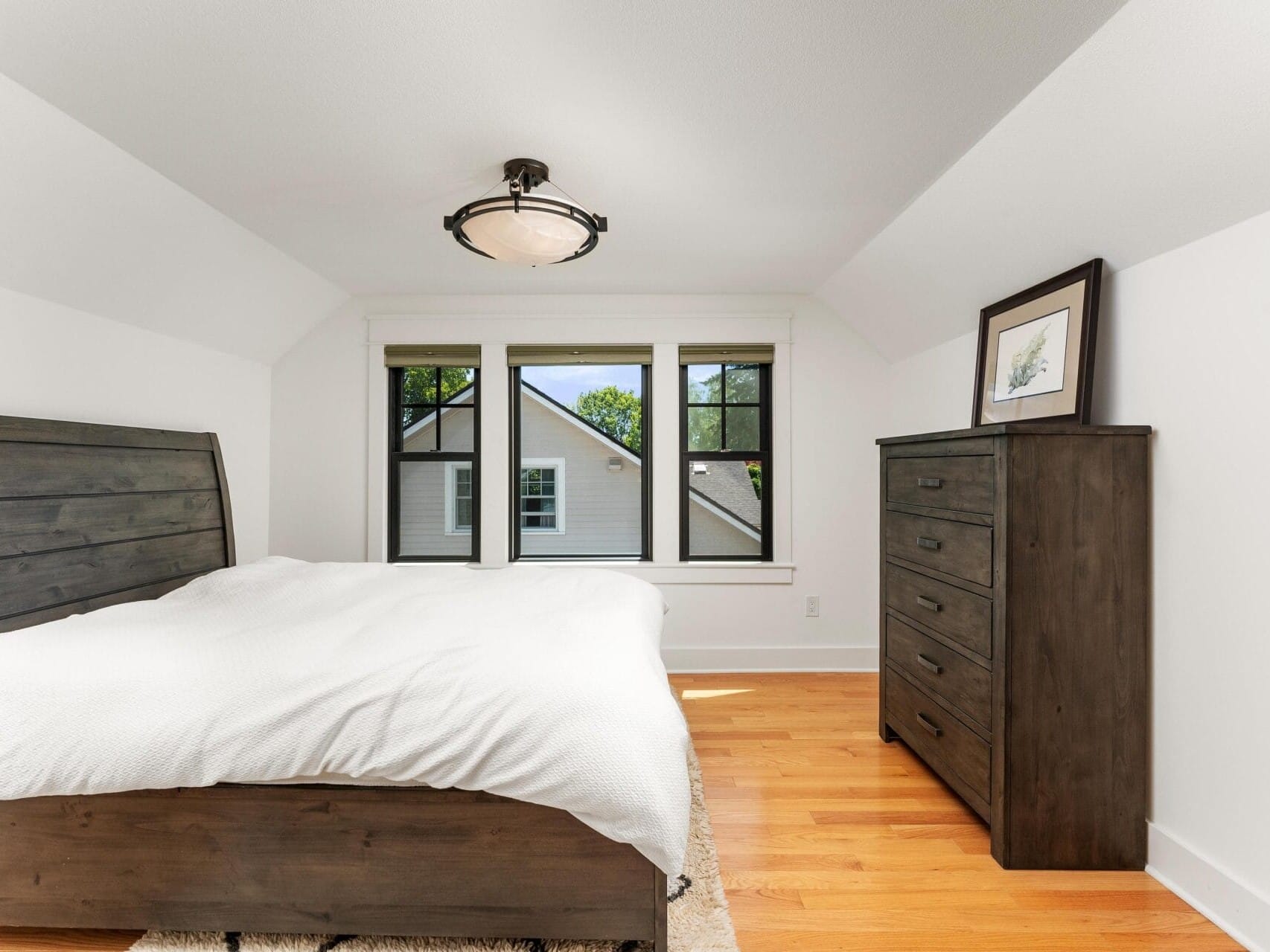 A minimalist bedroom with a wooden bed, white bedding, and a matching wooden dresser. Three windows are centered on the back wall, offering a view of a neighboring house. A ceiling light fixture hangs above the room with hardwood floors.