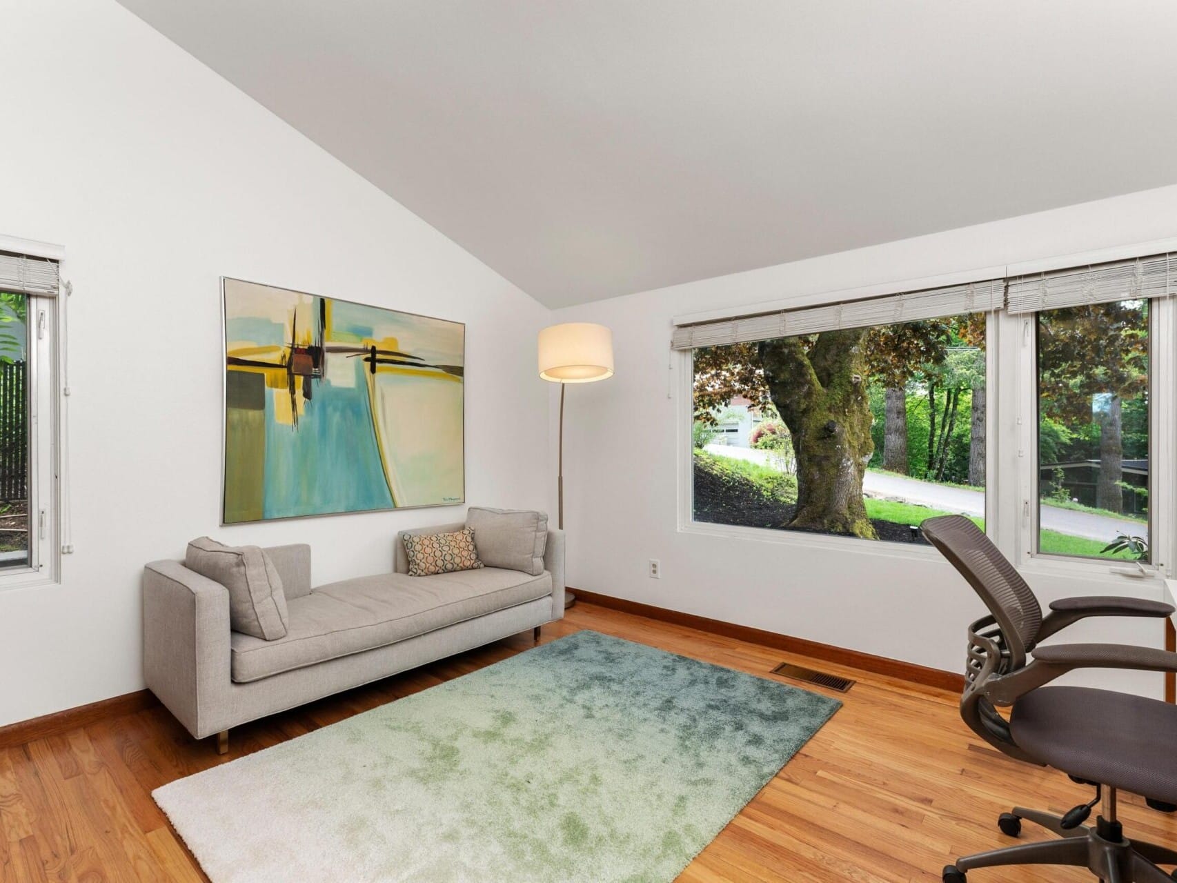 A modern living room with a light gray sofa, abstract painting, tall floor lamp, and two office chairs at a white desk. A large window looks out to a green garden. The floor is wooden with a gradient green rug.