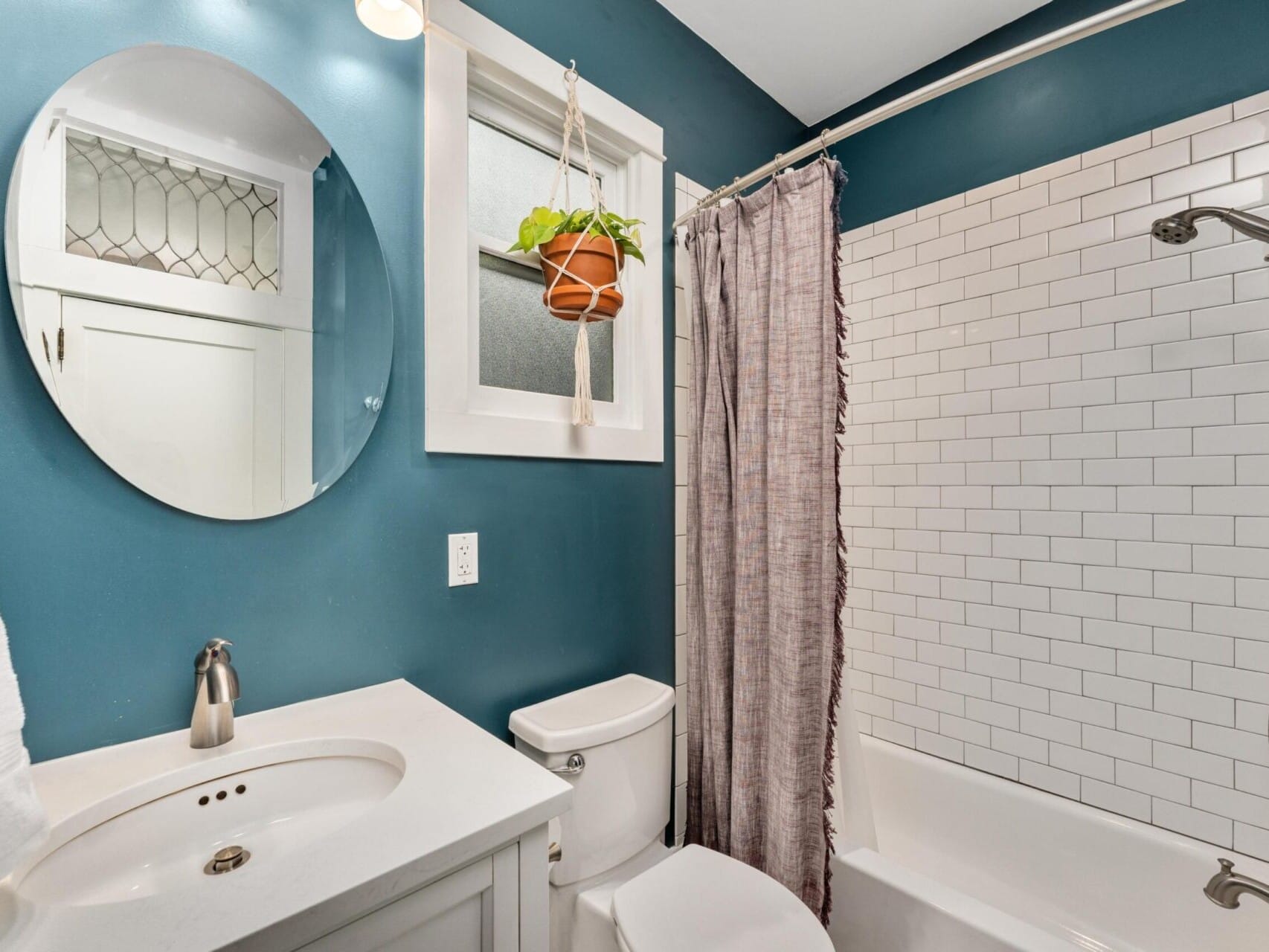 A small bathroom with blue walls features a round mirror above a white sink, a white toilet, and a bathtub with a subway tile shower. A plant hangs near a window above the toilet. The shower curtain is closed.