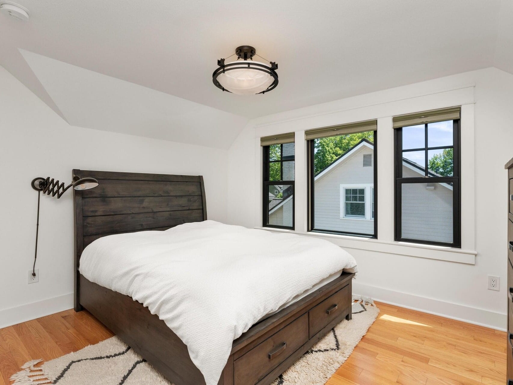 A cozy bedroom featuring a dark wooden bed with white bedding, a matching nightstand, a large window showing a neighboring house, and a modern ceiling light fixture. The room has wooden floors and white walls.