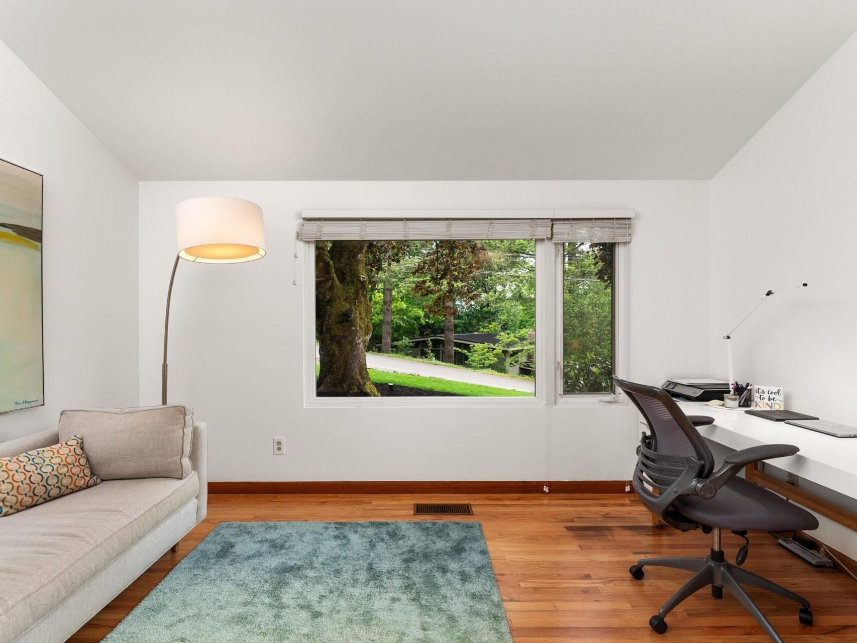 A modern home office with a large window showing greenery. Theres a gray sofa with a patterned pillow on the left, a floor lamp, and a desk with a chair and a computer on the right. The room has wooden floors and a light blue rug.