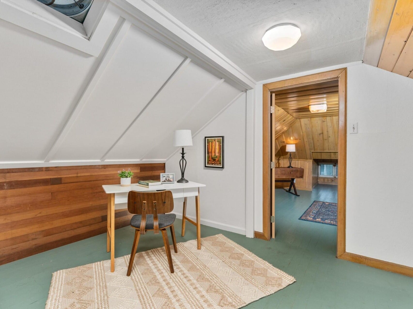 A cozy attic room with a sloped ceiling. It features a small white desk with a chair, a table lamp, and a framed picture. A doorway leads to another area, with wood paneling and a carpet visible. A rug covers part of the green floor.