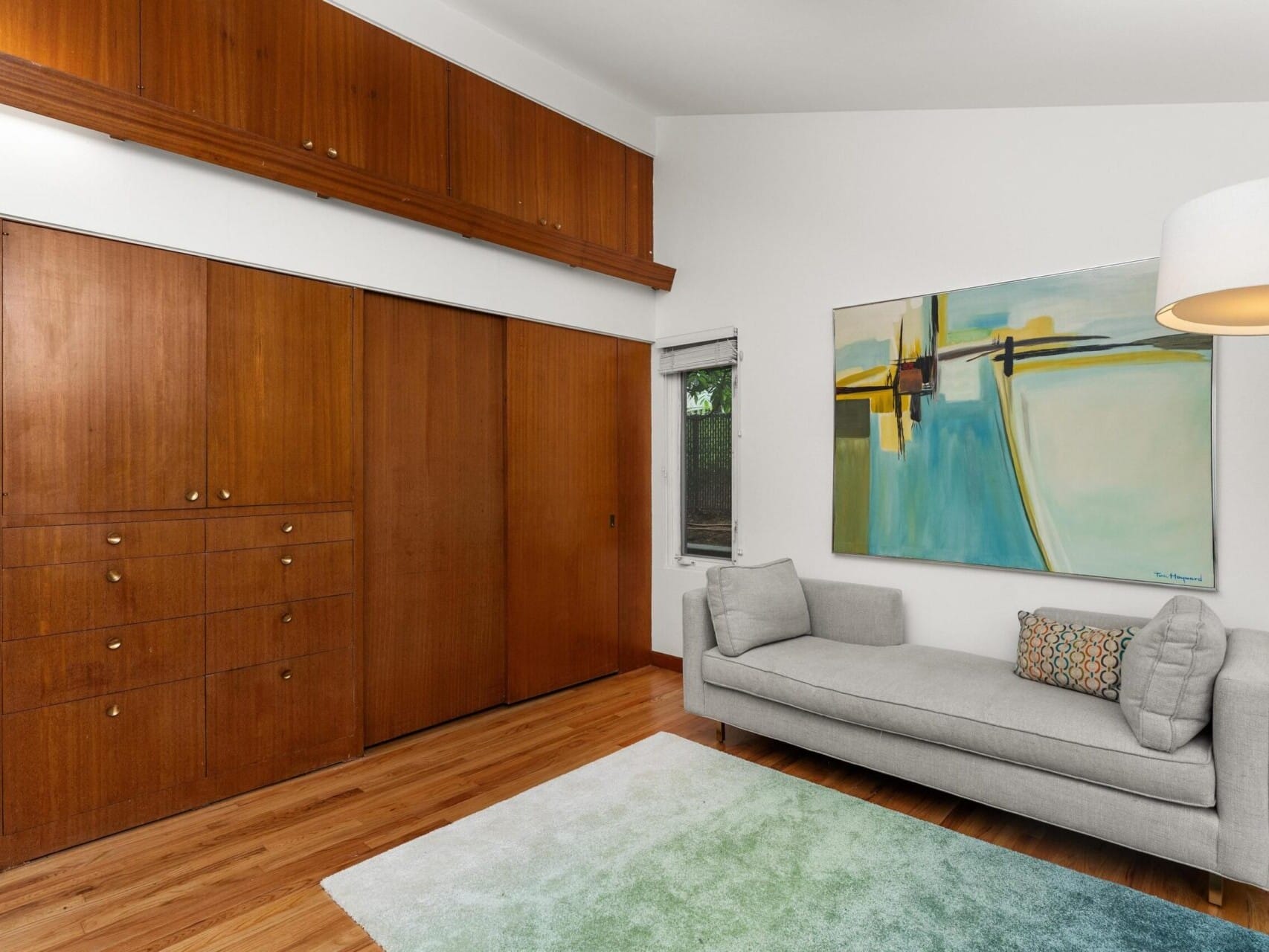 A modern living room features a gray chaise lounge with a decorative pillow, a colorful abstract painting on the wall, and wooden built-in cabinets. A soft, two-toned rug lies on the wooden floor, and a large white lamp provides lighting.