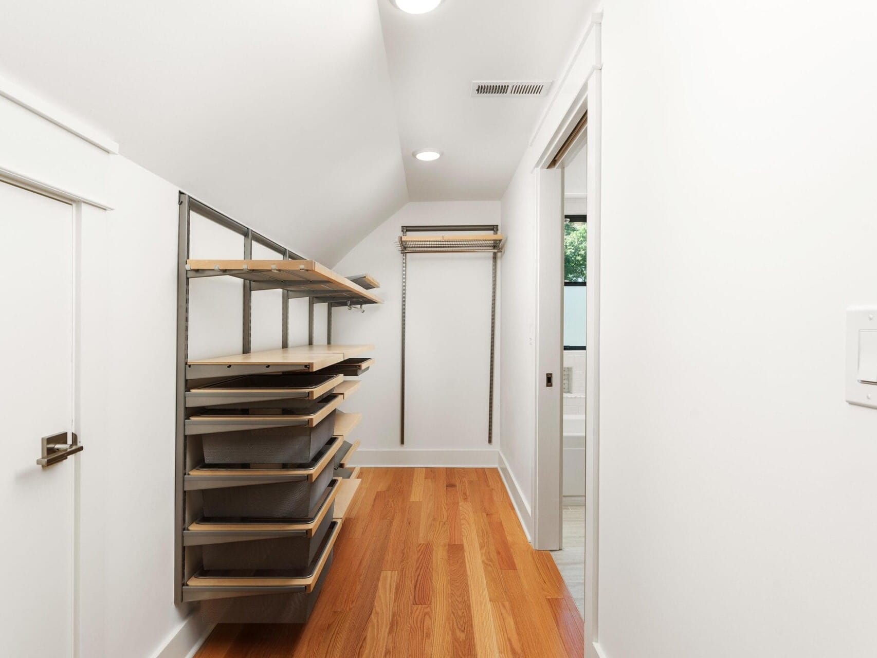 A narrow walk-in closet with wooden flooring and white walls. It features metal shelves and a hanging rod on the left side. A small door is on the left wall, and a bathroom is visible through an open door at the end.