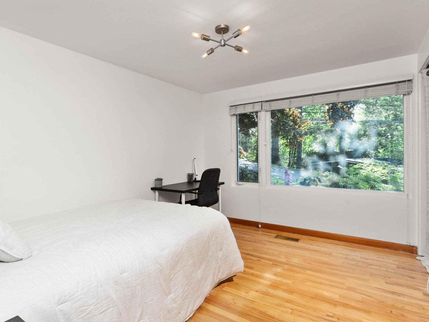 A minimalist bedroom with a wooden floor, white walls, and a large window showing greenery. The room features a bed with a white comforter, a black desk with a lamp and chair, and a modern ceiling light fixture. Light gray curtains frame the window.