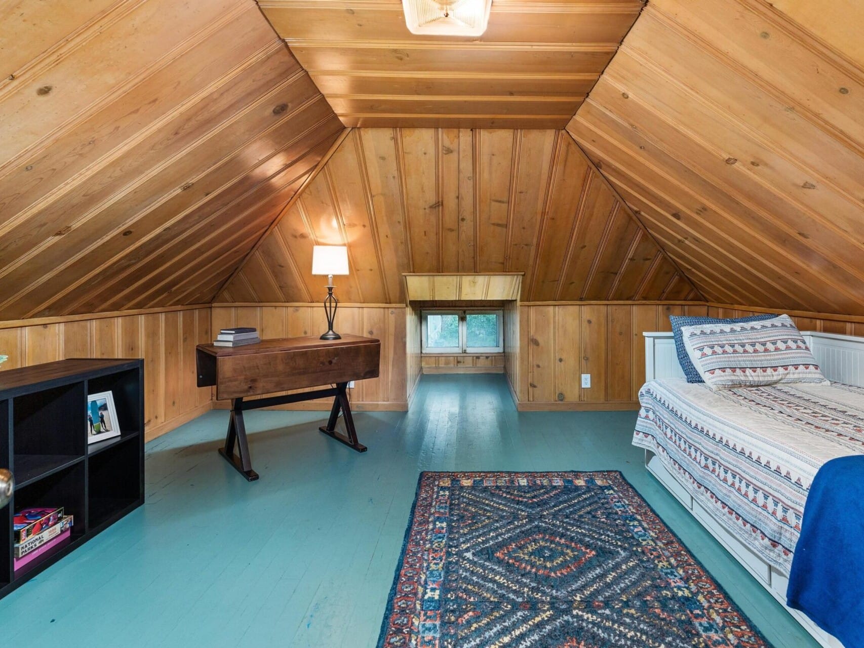 Attic room with sloped wooden ceiling, featuring a bed with patterned bedding, a freestanding lamp, a rug, a small window, a bookshelf, and a writing desk. The floor is painted a light blue-green color.