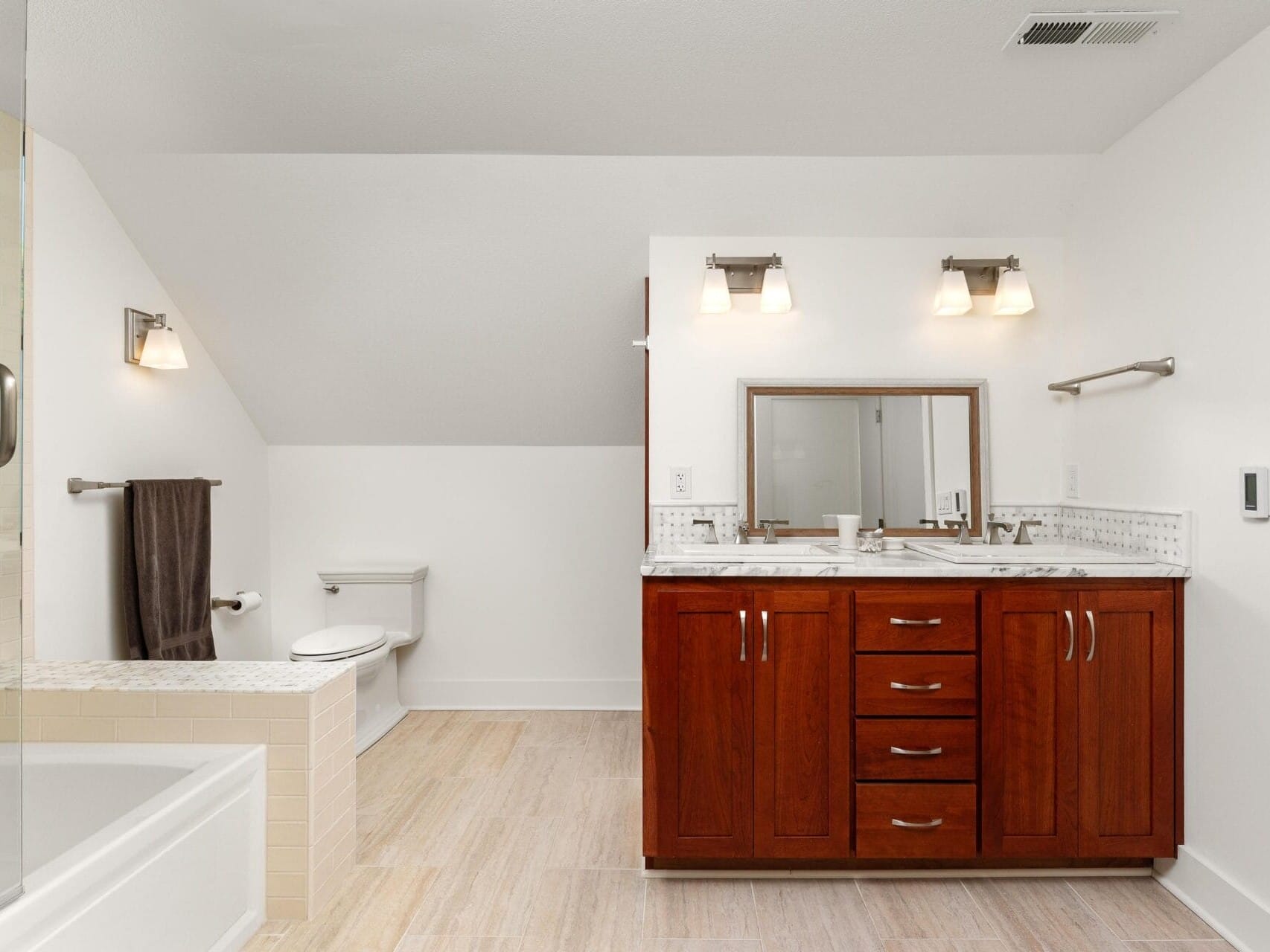 A modern bathroom with light wood floors, white walls, and brown wooden cabinets. It features a double-sink vanity with a large mirror, a bathtub, a toilet, and overhead lighting.