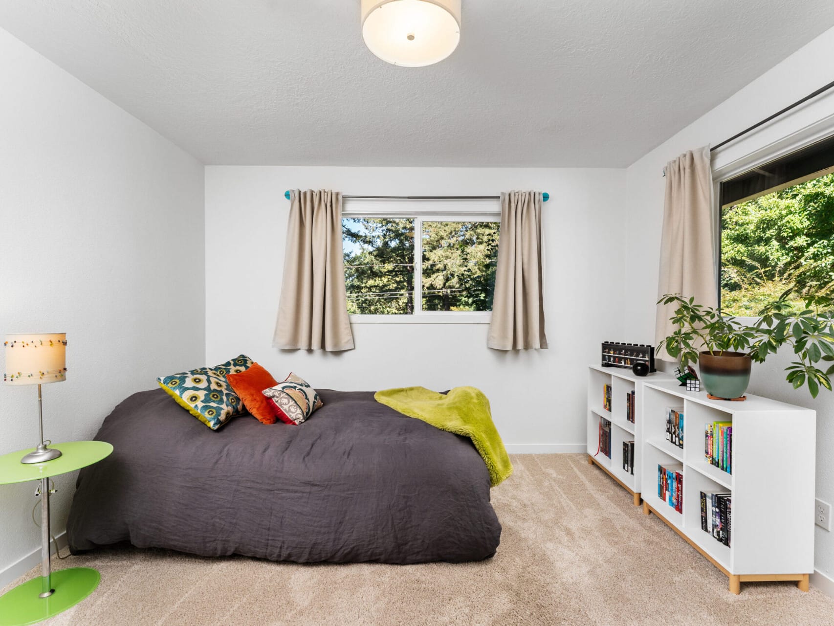 A cozy bedroom with a large gray bed, colorful pillows, and a green throw. A small round table with a decorative lamp stands beside it. There are two windows with beige curtains and a bookshelf filled with books and plants.