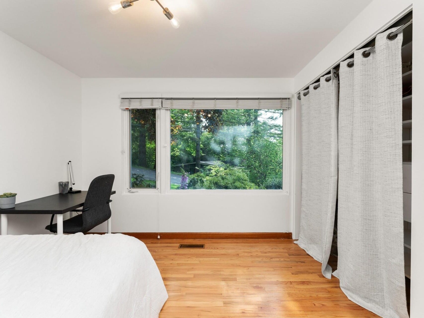 A minimalist bedroom with a hardwood floor, featuring a bed with a white comforter, a small black desk with a chair by a large window offering a view of greenery, and a curtain covering a closet. A framed picture hangs on the right wall.