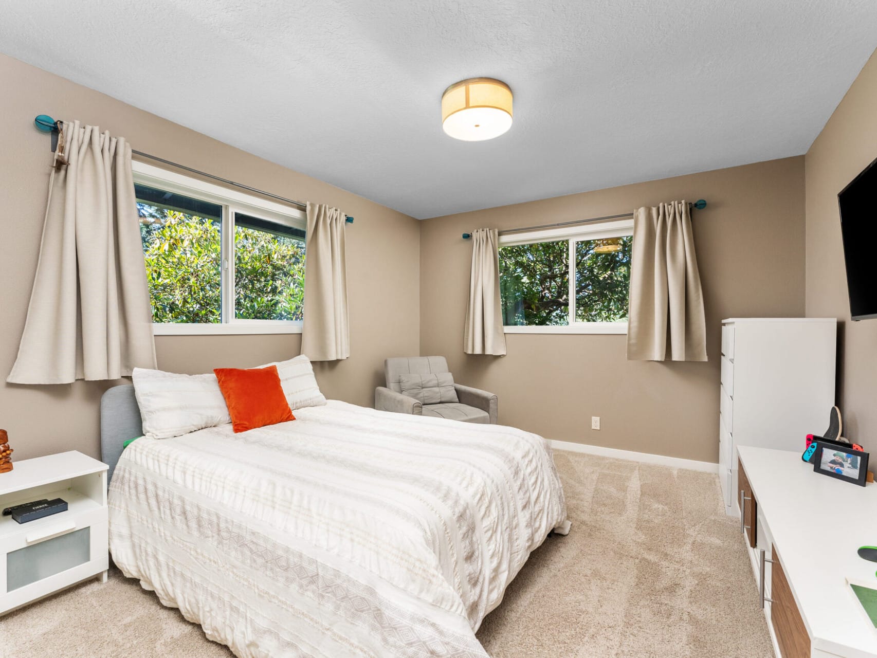 A modern bedroom with beige walls features a double bed with a white comforter and orange accent pillow. Two windows with curtains allow natural light. Theres a gray armchair, a wall-mounted TV, and a white dresser. A ceiling light fixture is above.