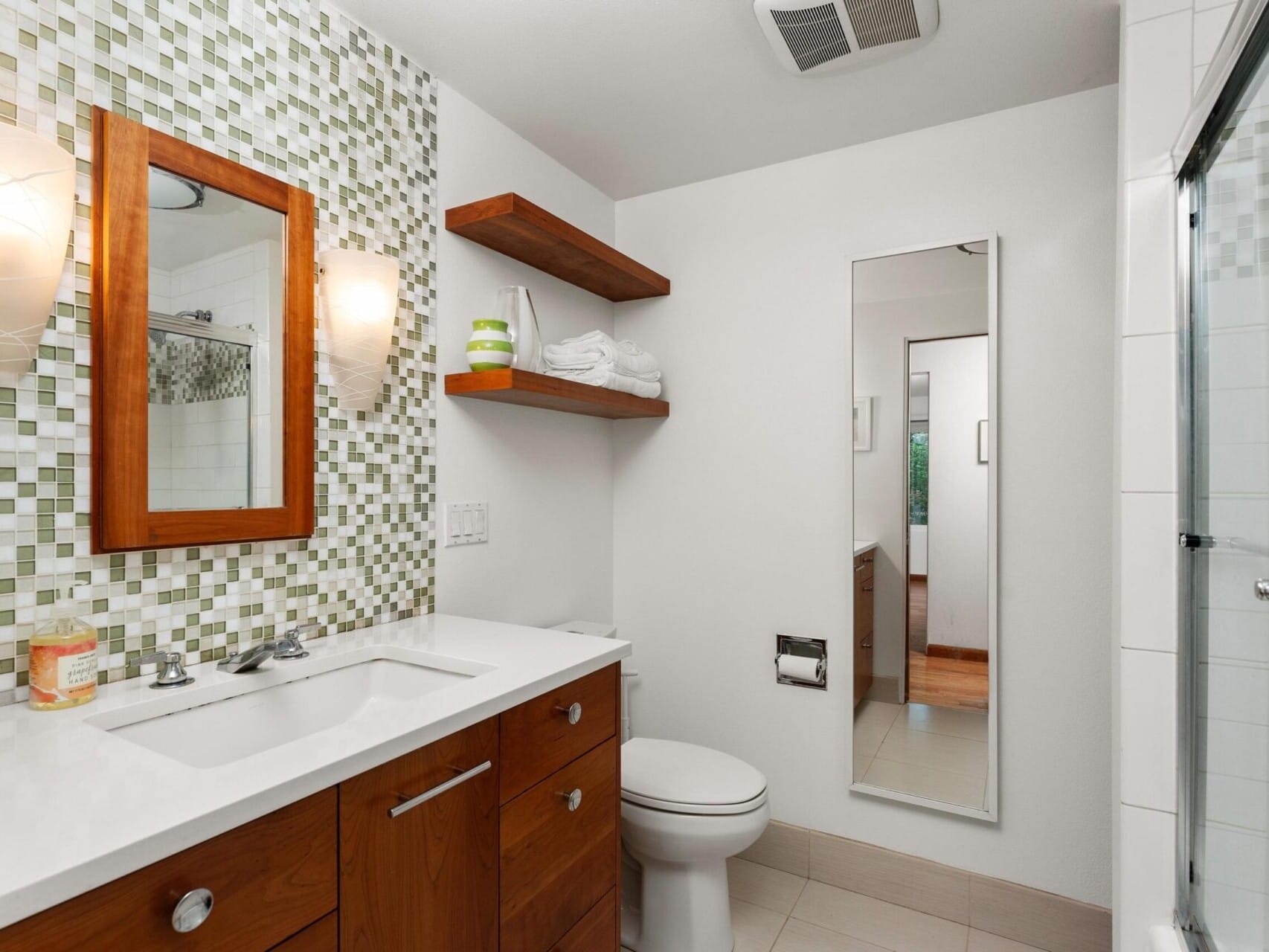 A modern bathroom with a wooden vanity and white countertop, mosaic tile backsplash, and two wall-mounted lights. There are open shelves with towels, a large mirror, and a glass-enclosed shower. The floor is tiled in a light beige color.