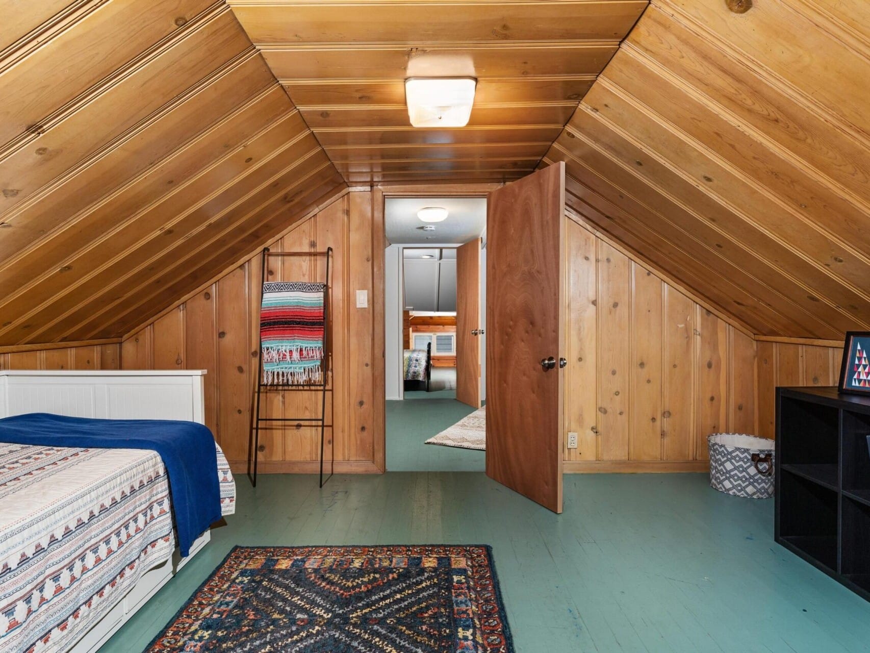 A cozy attic room with wooden paneling and sloped ceiling. It features a bed with a patterned blanket, a ladder rack with blankets, an open door leading to another room, and a bookshelf. A colorful rug lies on the painted wooden floor.