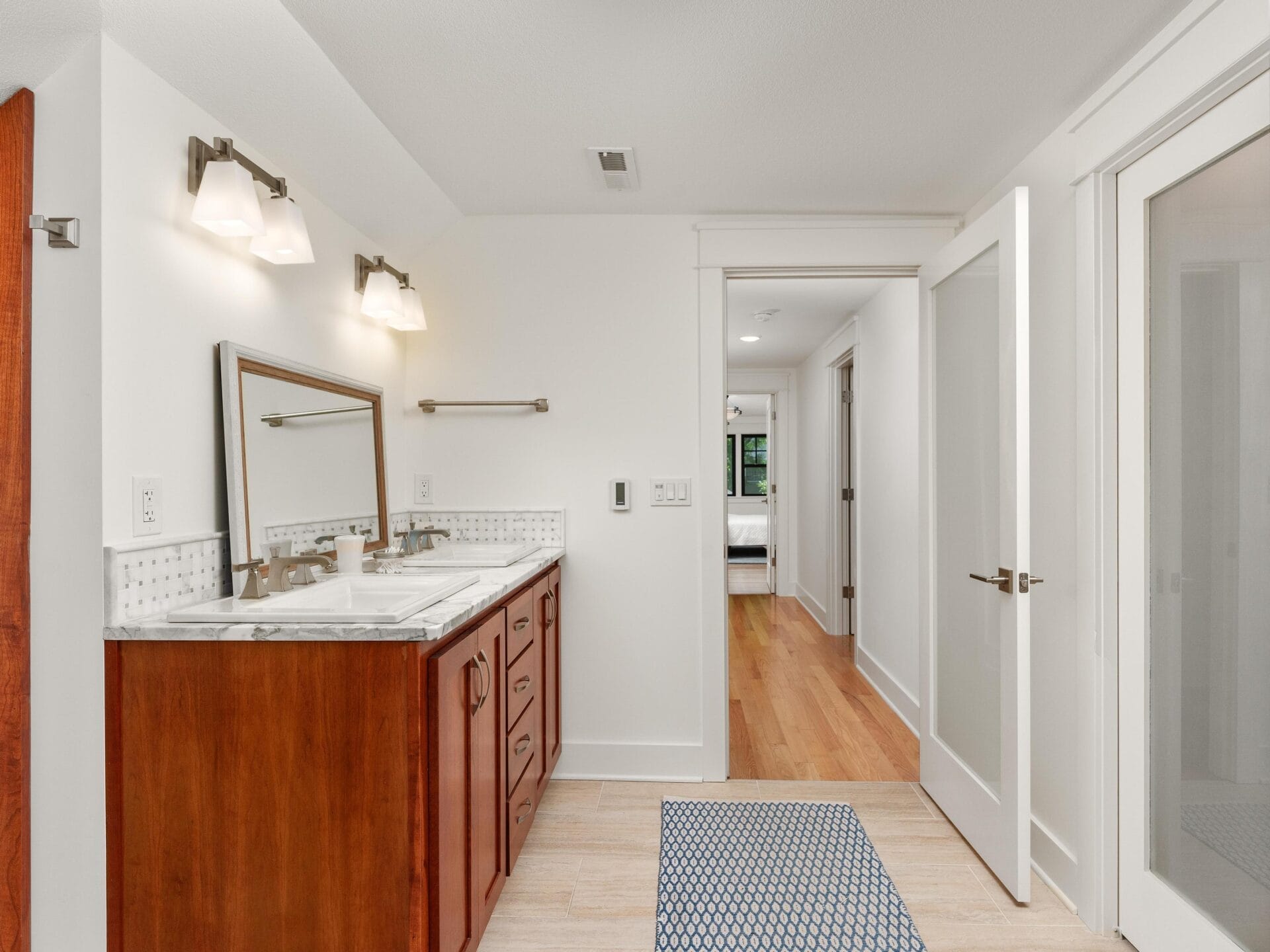 A modern bathroom with a double sink vanity featuring a marble countertop and wooden cabinets. Above the sinks are two wall-mounted lights. The floor is tiled, and a door leads to a hallway with wooden flooring. A small rug is placed near the sinks.