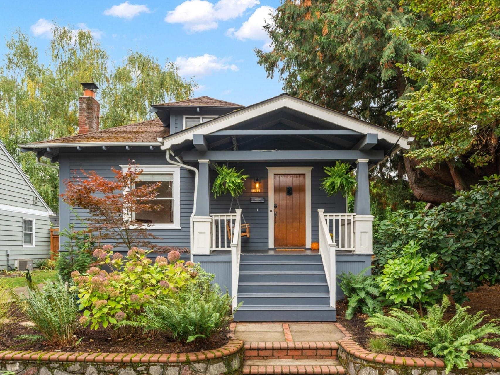 A charming blue house with a wooden front door, white trim, and a cozy porch. The yard features lush greenery, vibrant flowers, and large ferns. A brick path leads to the entrance. The sky is partly cloudy, with trees surrounding the house.