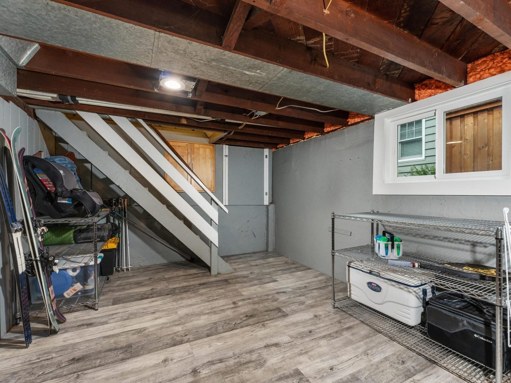 A basement with exposed wooden beams, gray walls, and wood-like flooring. It features a staircase, sports equipment, and a shelf with boxes and cleaning supplies. A small window lets in natural light.