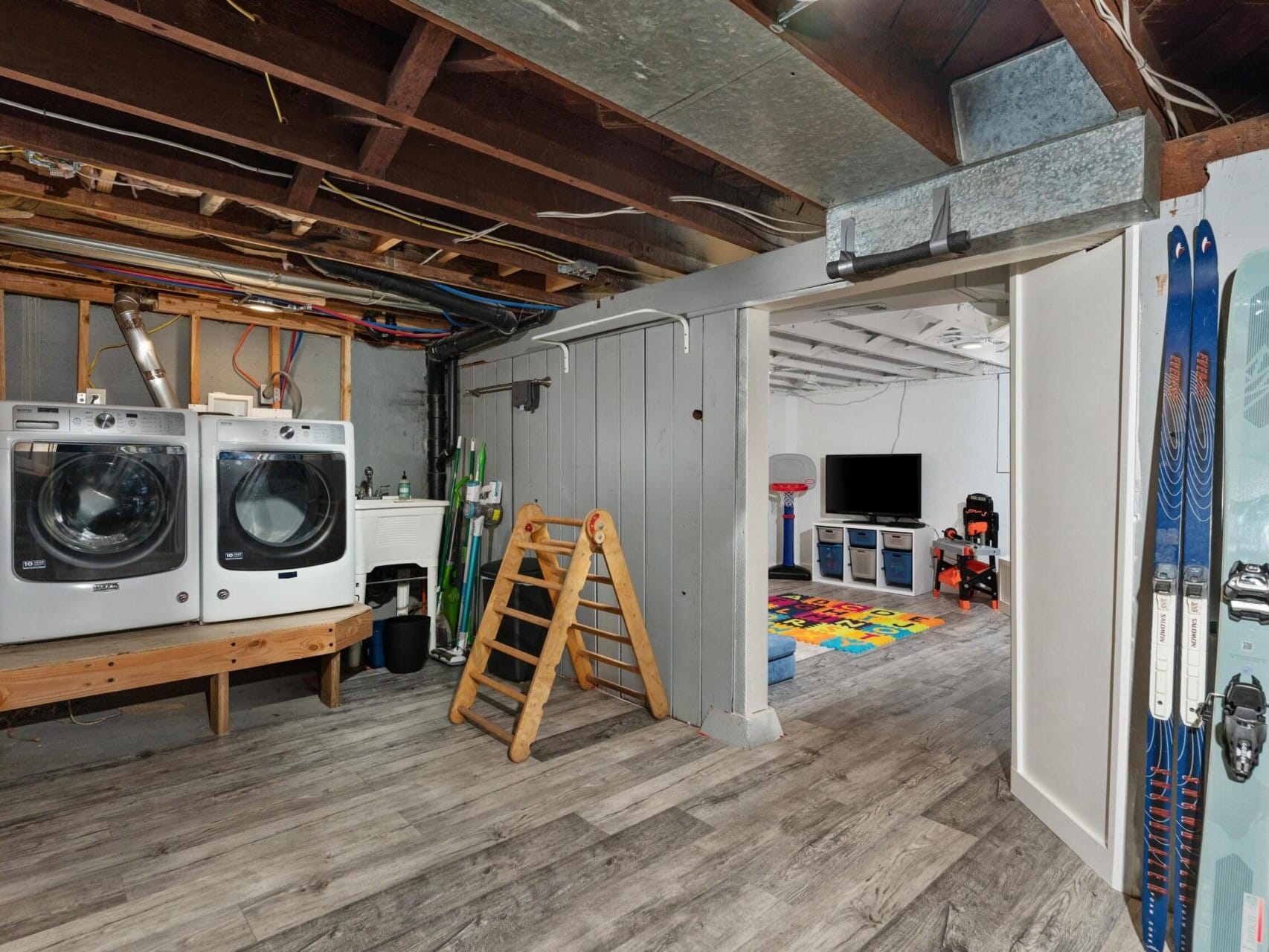 A basement with a laundry area featuring a washer and dryer, a sink, and cleaning supplies. To the right, theres a play area with colorful foam tiles, a TV, and toys. Skis lean against the wall. The ceiling shows exposed beams and ductwork.