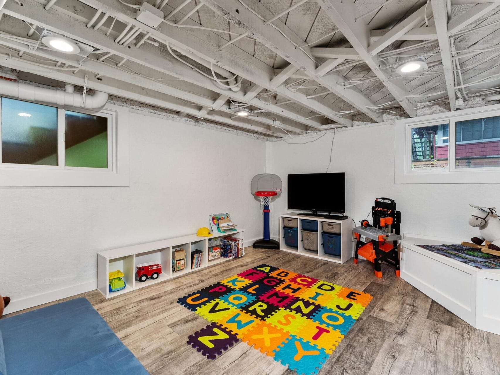 A playroom with exposed beams, featuring a colorful alphabet mat, small basketball hoop, TV, toy storage, and a ride-on toy. The walls are white, and the floor is wooden. Natural light comes through two windows.