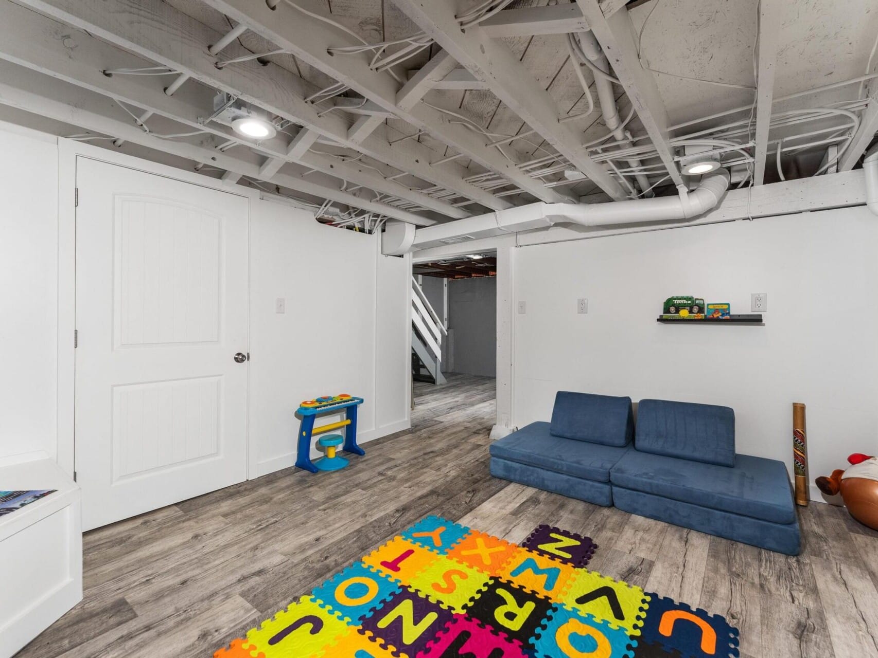 A basement playroom with exposed ceiling beams, featuring a blue couch, colorful alphabet play mat on the wooden floor, and a small blue play kitchen. Toys are visible on a shelf, and a door leads to another room.