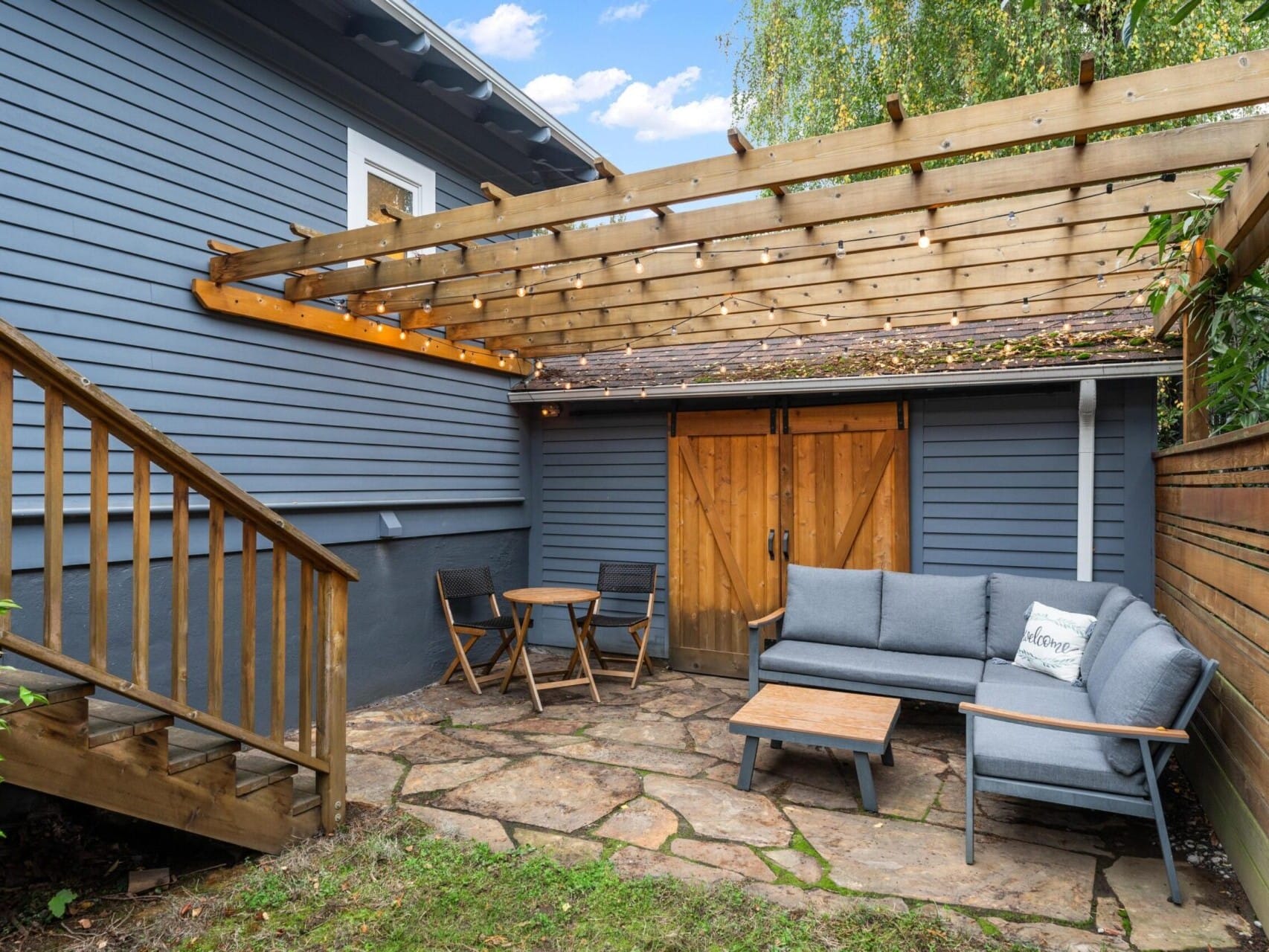 A cozy patio with a wooden pergola, featuring a gray sectional sofa, a wooden coffee table, and a small round table with two chairs. The area is enclosed by wooden fencing and bordered by green shrubs, with a stone tile floor.