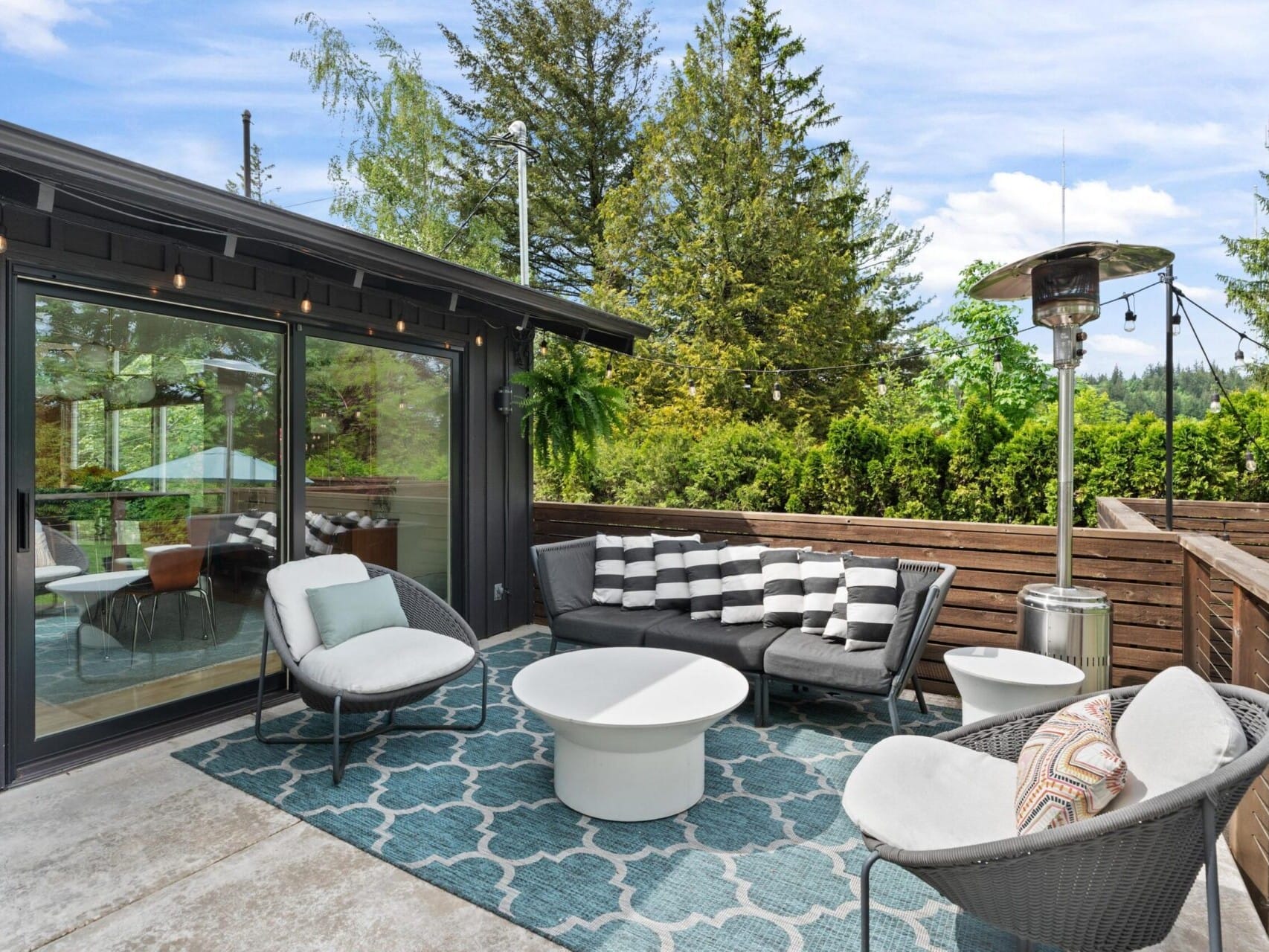 A cozy outdoor patio with modern furniture, including a gray sofa, two round chairs, and a circular white table. A blue geometric rug covers the floor. String lights and a patio heater add warmth, surrounded by lush trees and a cloudy sky.