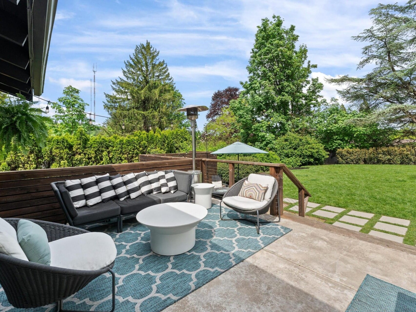 A modern outdoor patio with a comfortable seating area featuring a black and white cushioned sofa, two round white tables, and chairs on a patterned blue rug. A patio heater stands nearby, and a manicured lawn and trees are in the background.