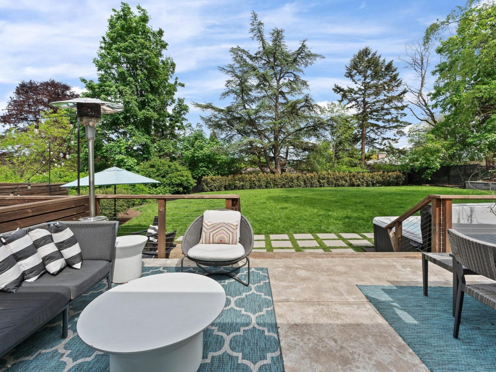 A spacious outdoor patio features modern gray furniture, a round coffee table, and patterned rugs. It overlooks a lush green lawn with tall trees and a blue sky in the background. Strings of lights hang across the space, adding a cozy touch.