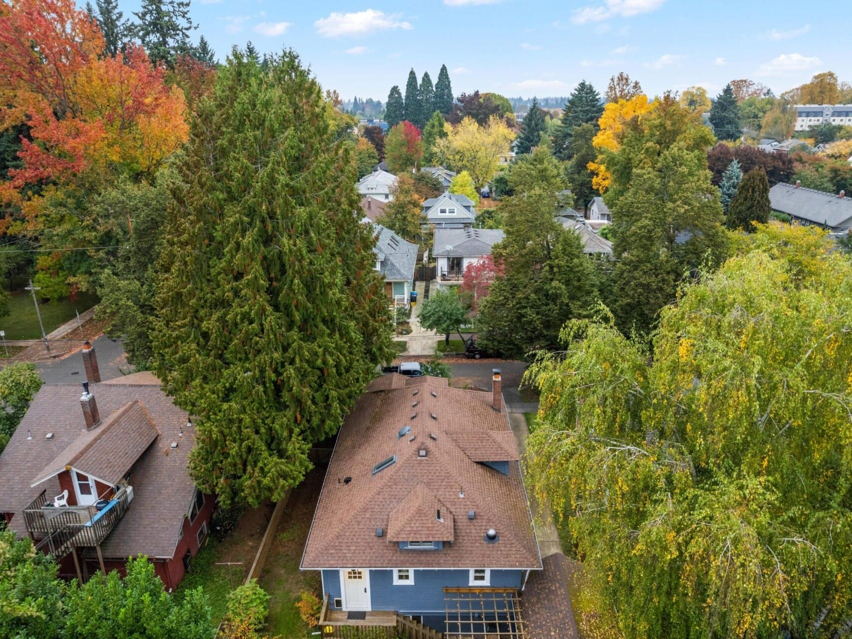 Aerial view of a residential neighborhood in autumn, featuring houses surrounded by tall trees with vibrant fall foliage in shades of green, orange, and yellow. The sky is clear with a few clouds.