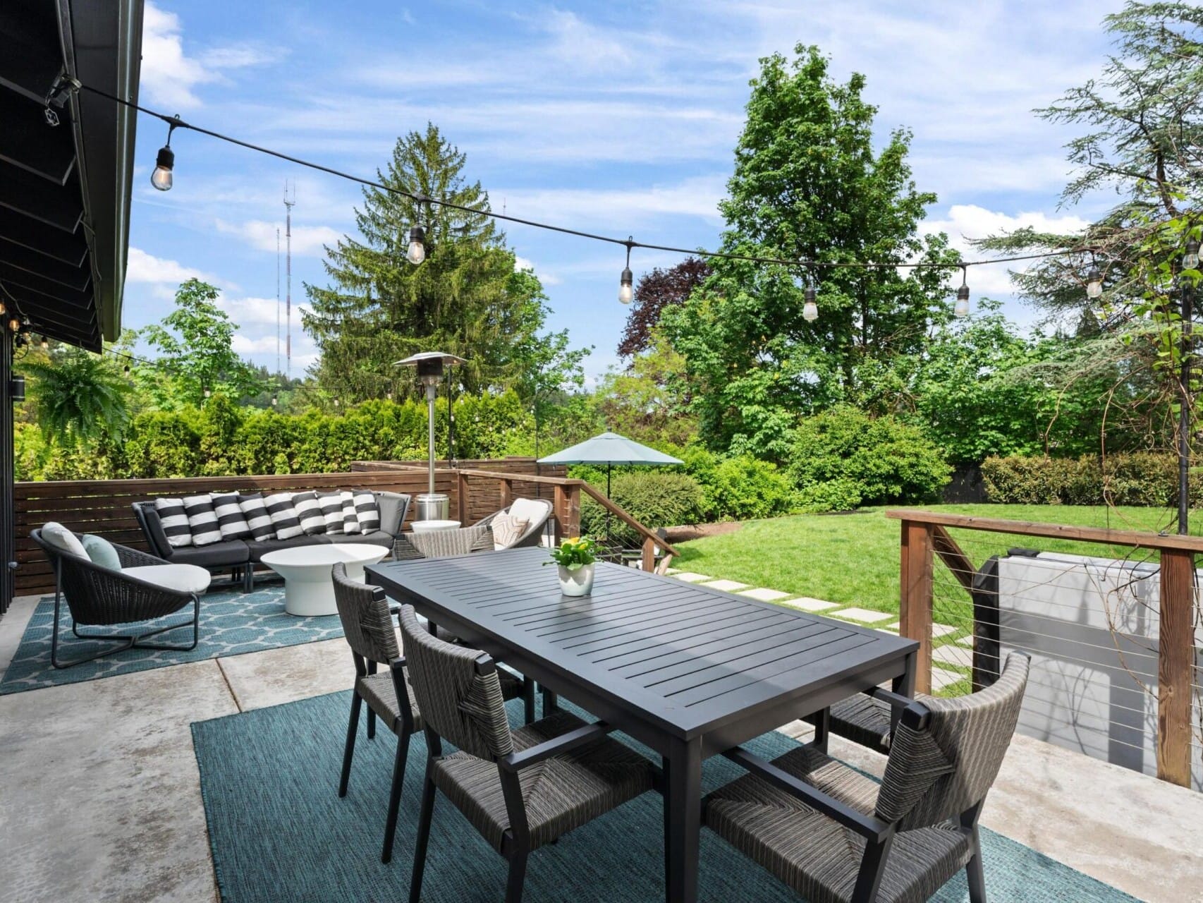 A spacious outdoor patio featuring a long dining table with chairs and a modern seating area under string lights. The patio overlooks a lush green lawn surrounded by trees, with a sunny, blue sky in the background.