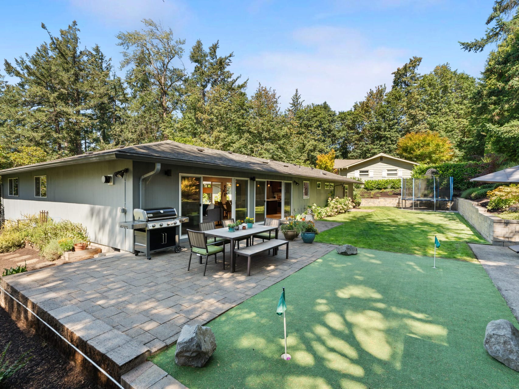 A backyard features a putting green with several holes, a stone patio with a BBQ grill, a dining table, and a trampoline. The area is surrounded by trees, and a single-story house is visible in the background.