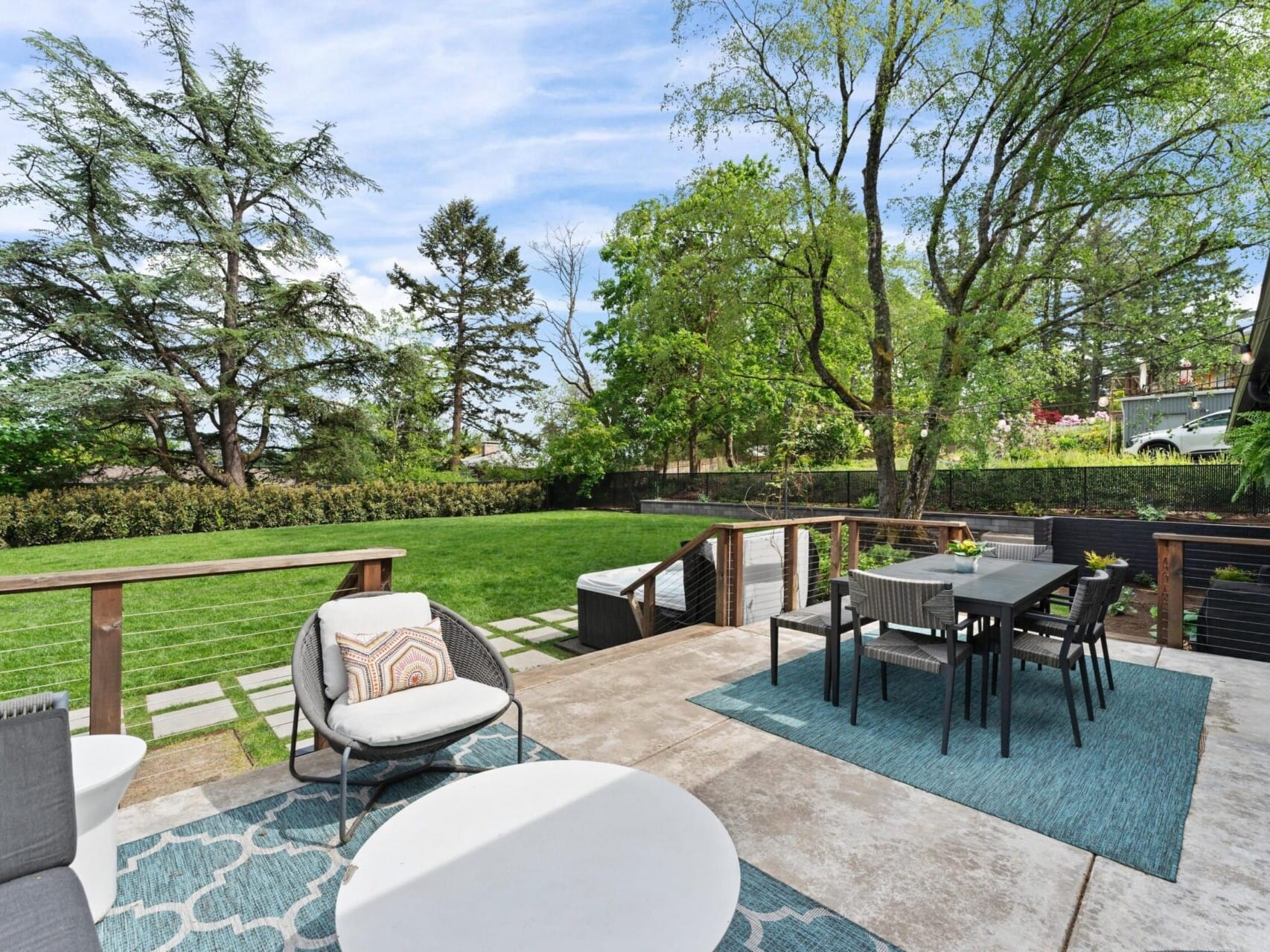 A modern outdoor patio with a dining table and chairs on a blue rug, surrounded by a spacious lawn and trees. The patio has a circular coffee table and chairs with cushions, and a railing separates the patio from the grass.