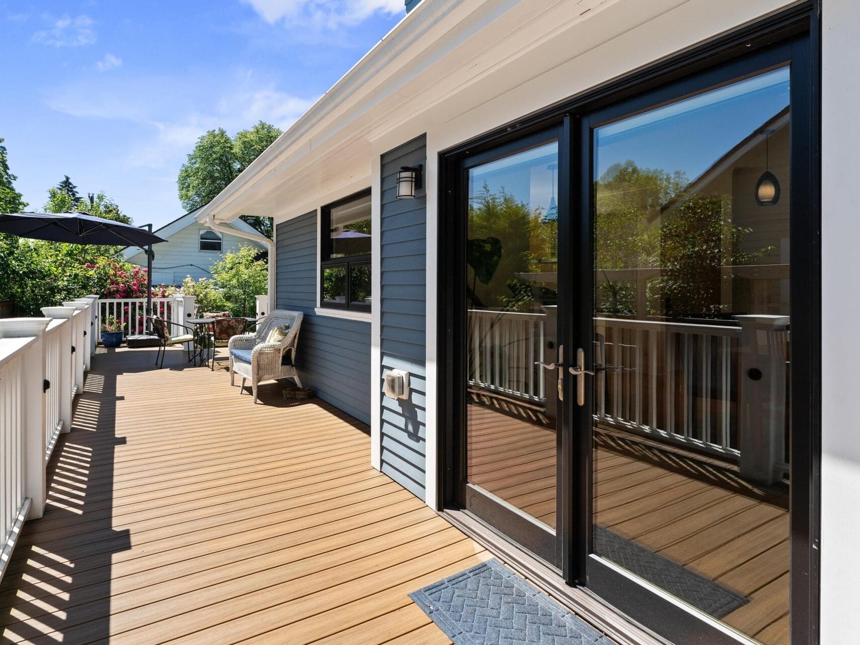 A wooden deck with railings attached to a blue house. Glass double doors reflect the outdoors. Theres outdoor seating, including a wicker chair and table with an umbrella. Trees and neighboring houses are visible under a clear blue sky.