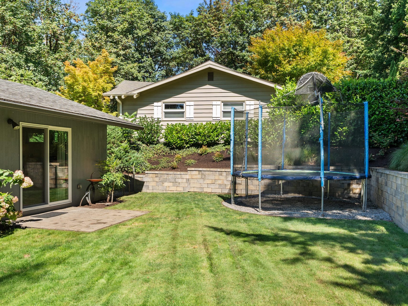 A backyard with a well-kept lawn, a trampoline with a safety net, surrounded by greenery and trees. A beige house with a small patio is on the left, and a stone retaining wall separates the garden areas.