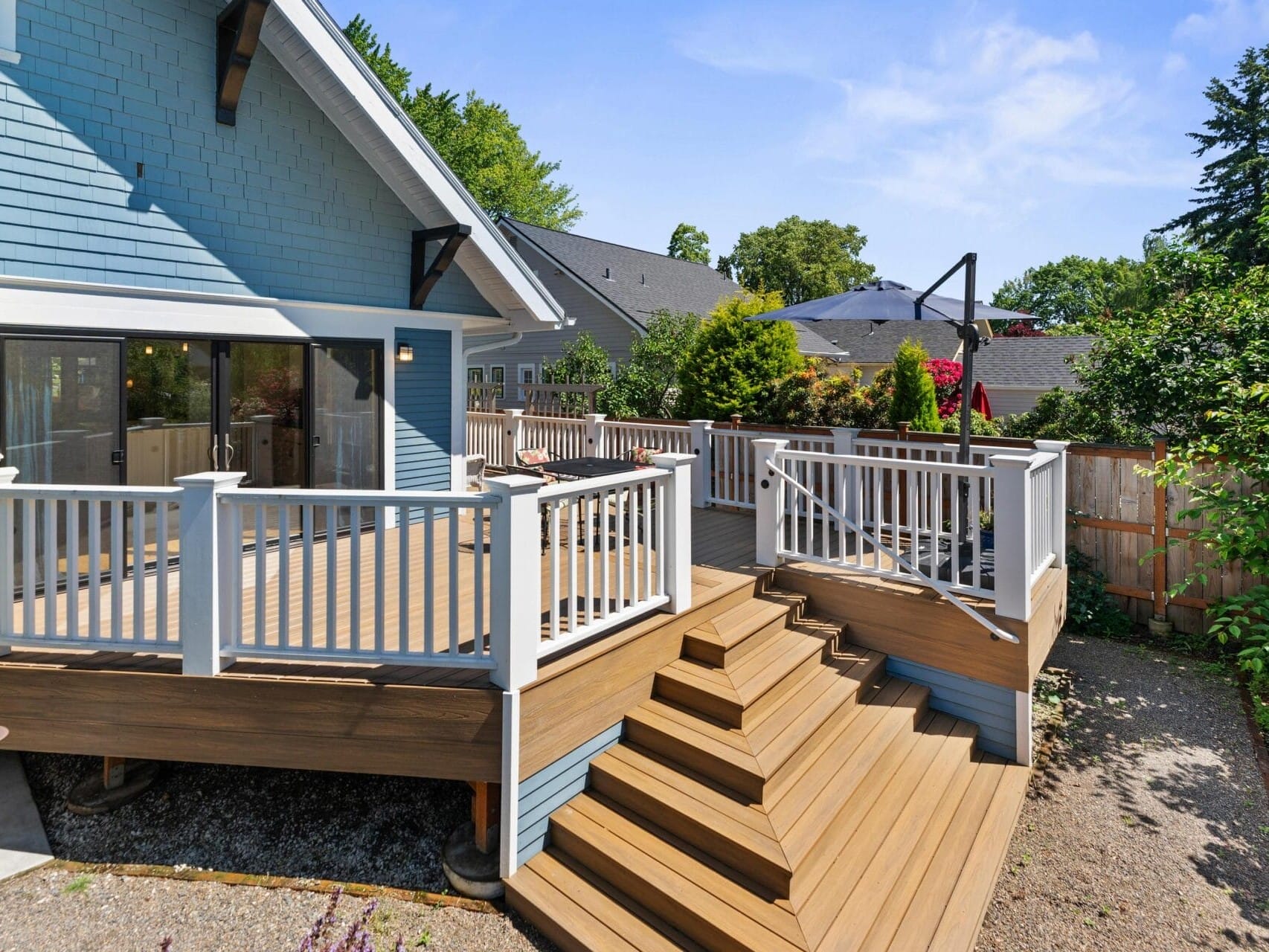 A modern backyard with a spacious wooden deck featuring white railings and steps. The house is light blue with large windows and a sloped roof. Surrounding trees and a clear sky add to the serene atmosphere.