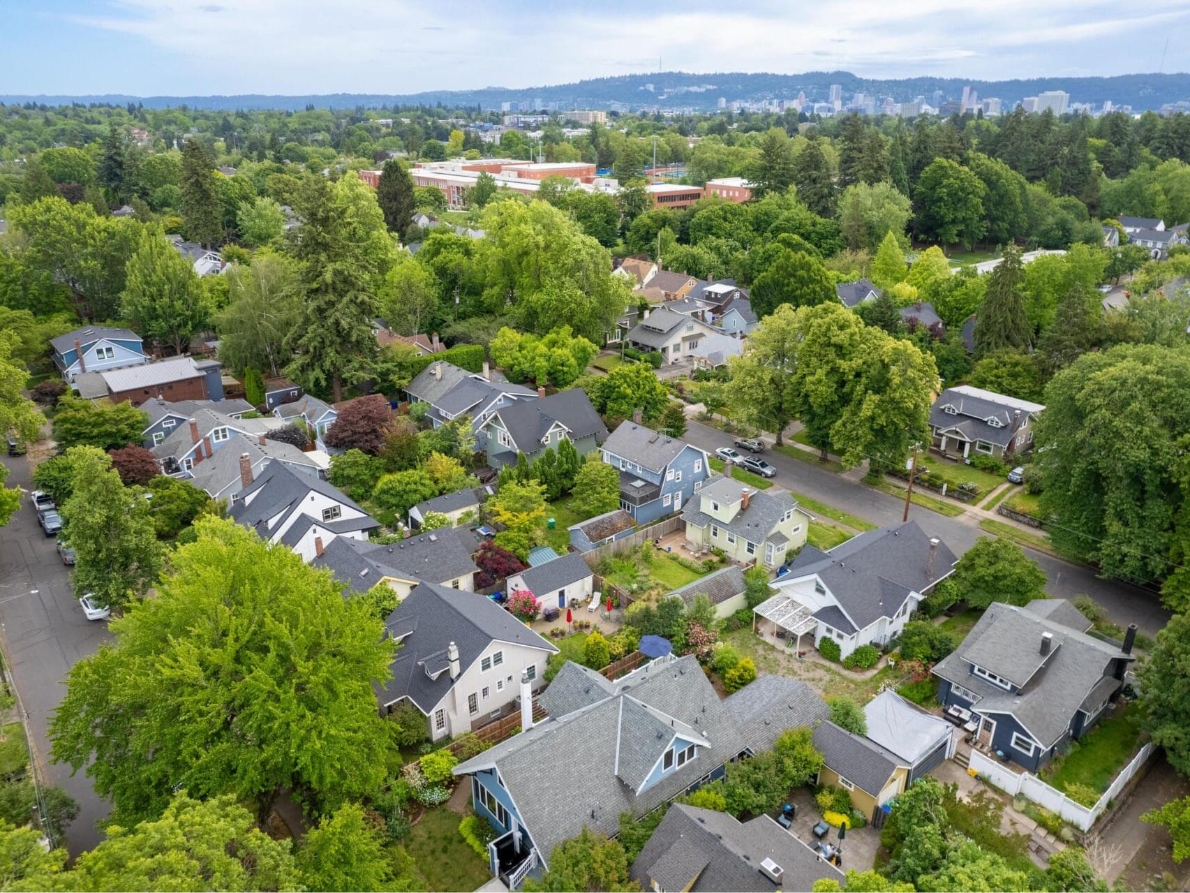 Aerial view of a suburban neighborhood with tree-lined streets and diverse houses. Lush greenery surrounds the residential area, with a city skyline visible in the distance under a cloudy sky.