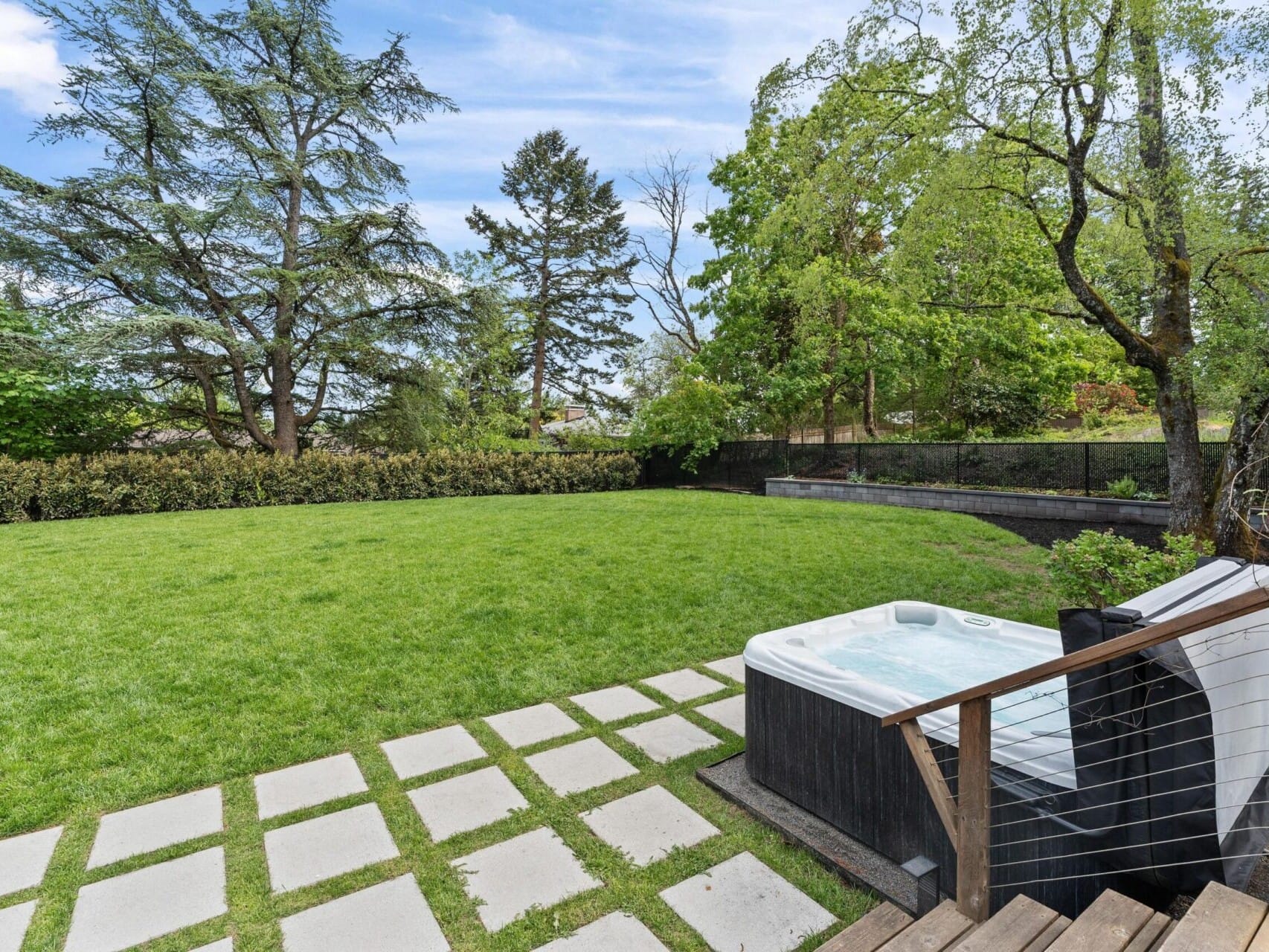 A grassy backyard with a paved pathway leading to a wooden deck and hot tub. Surrounding the area are tall trees and a hedge, creating a private and serene outdoor space under a clear blue sky.