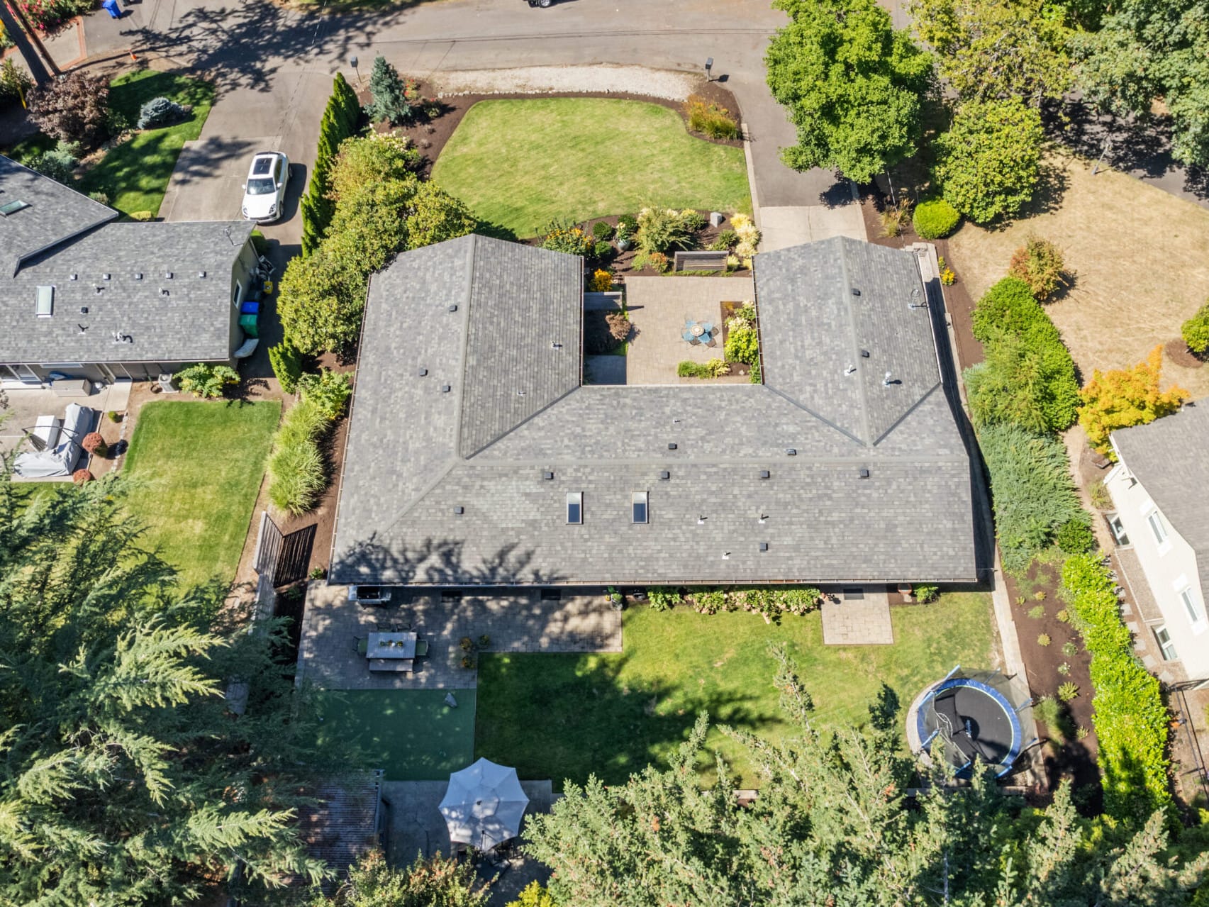 Aerial view of a suburban neighborhood featuring a large, U-shaped house with a gray roof. The property includes a green lawn, surrounding trees, a trampoline, and a small gazebo. Adjacent homes and a road are visible.