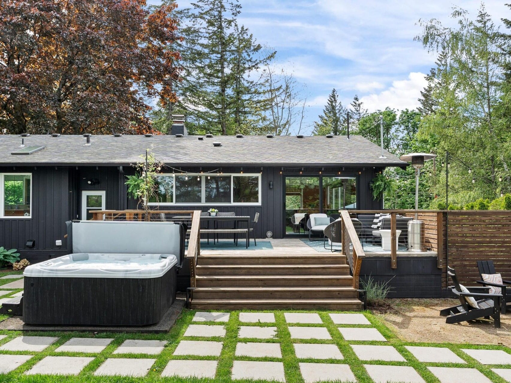 A modern backyard featuring a black-painted house with a wooden deck. The yard has a hot tub, a dining area with chairs, and a fire pit area with Adirondack chairs. The landscape includes stepping stones, lush greenery, and tall trees.