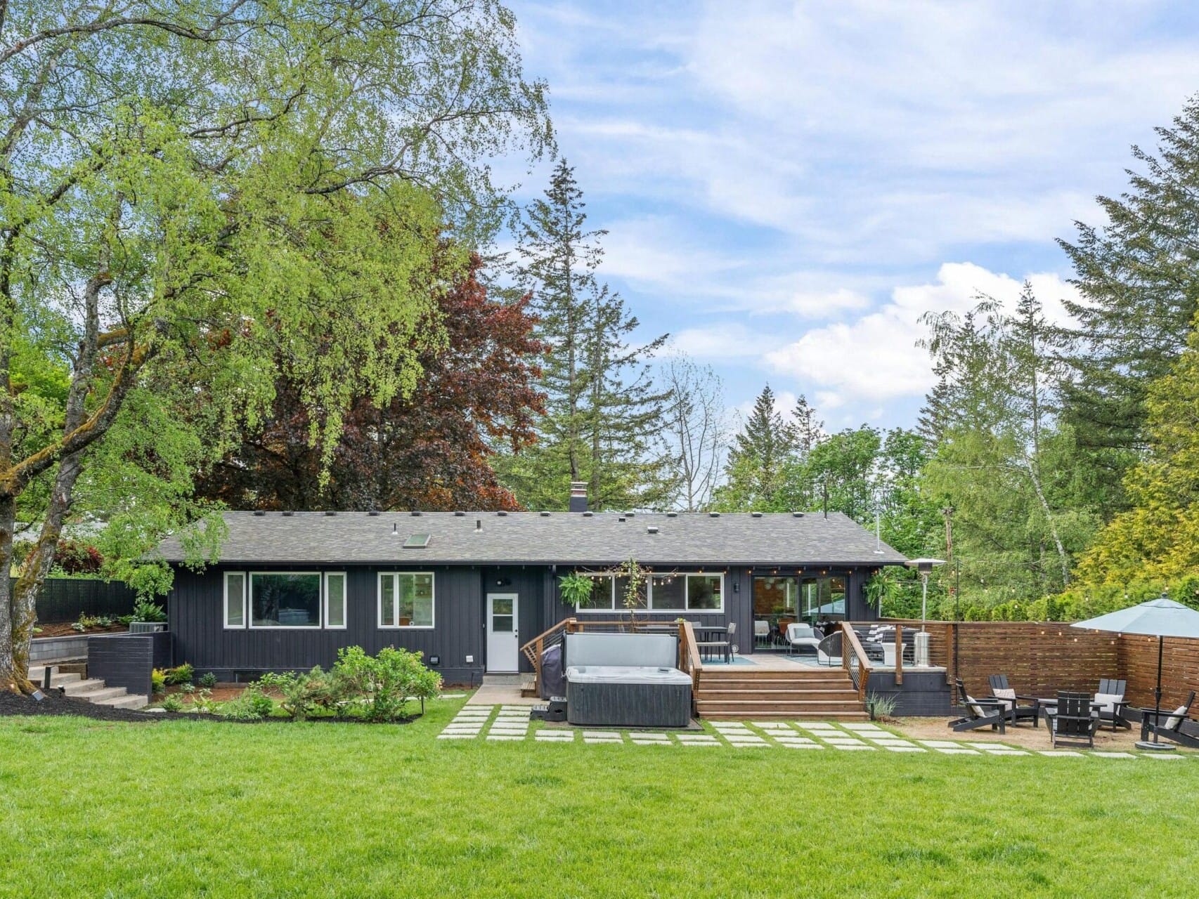 A modern house with dark siding, surrounded by lush green trees and a lawn. In the backyard, theres a wooden deck, a hot tub, a patio with chairs, and a sunshade. The sky is partly cloudy.