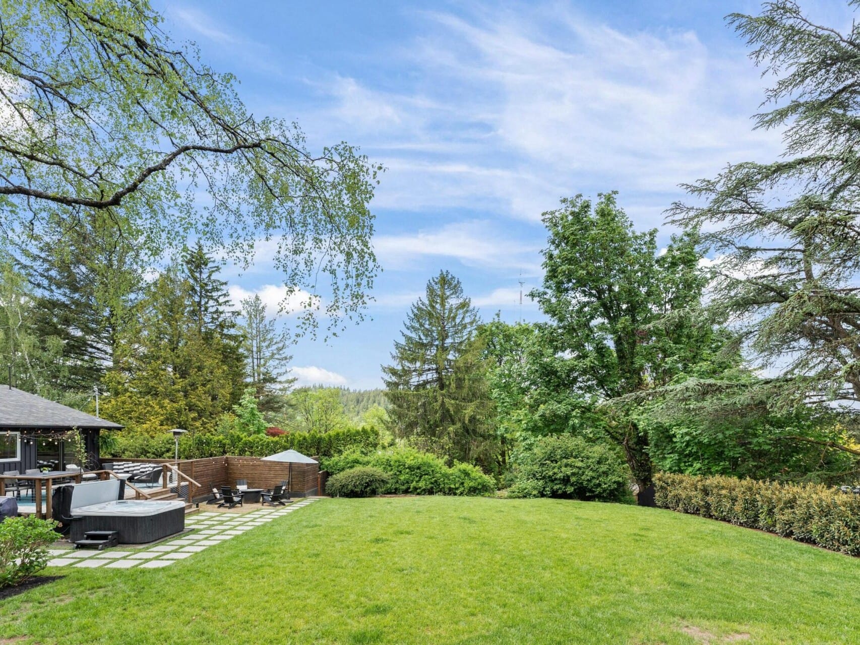 Spacious backyard with a manicured lawn, bordered by tall trees and bushes. Theres a wooden deck on the left side, featuring a hot tub and outdoor seating area under a canopy, against a backdrop of a partly cloudy sky.